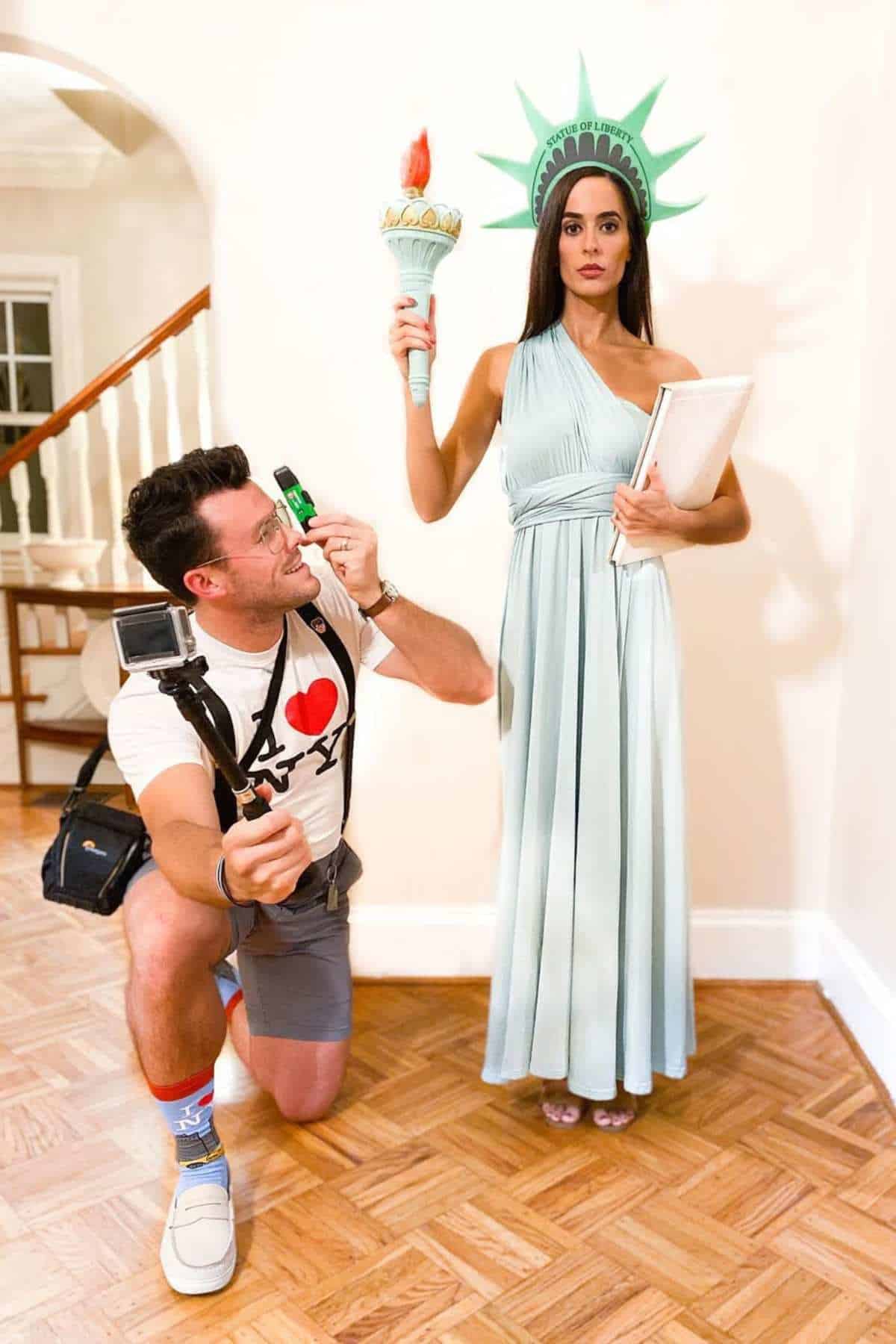 A man in tourist attire kneels with a camera and map, posing next to a woman dressed as the Statue of Liberty holding a torch and book.