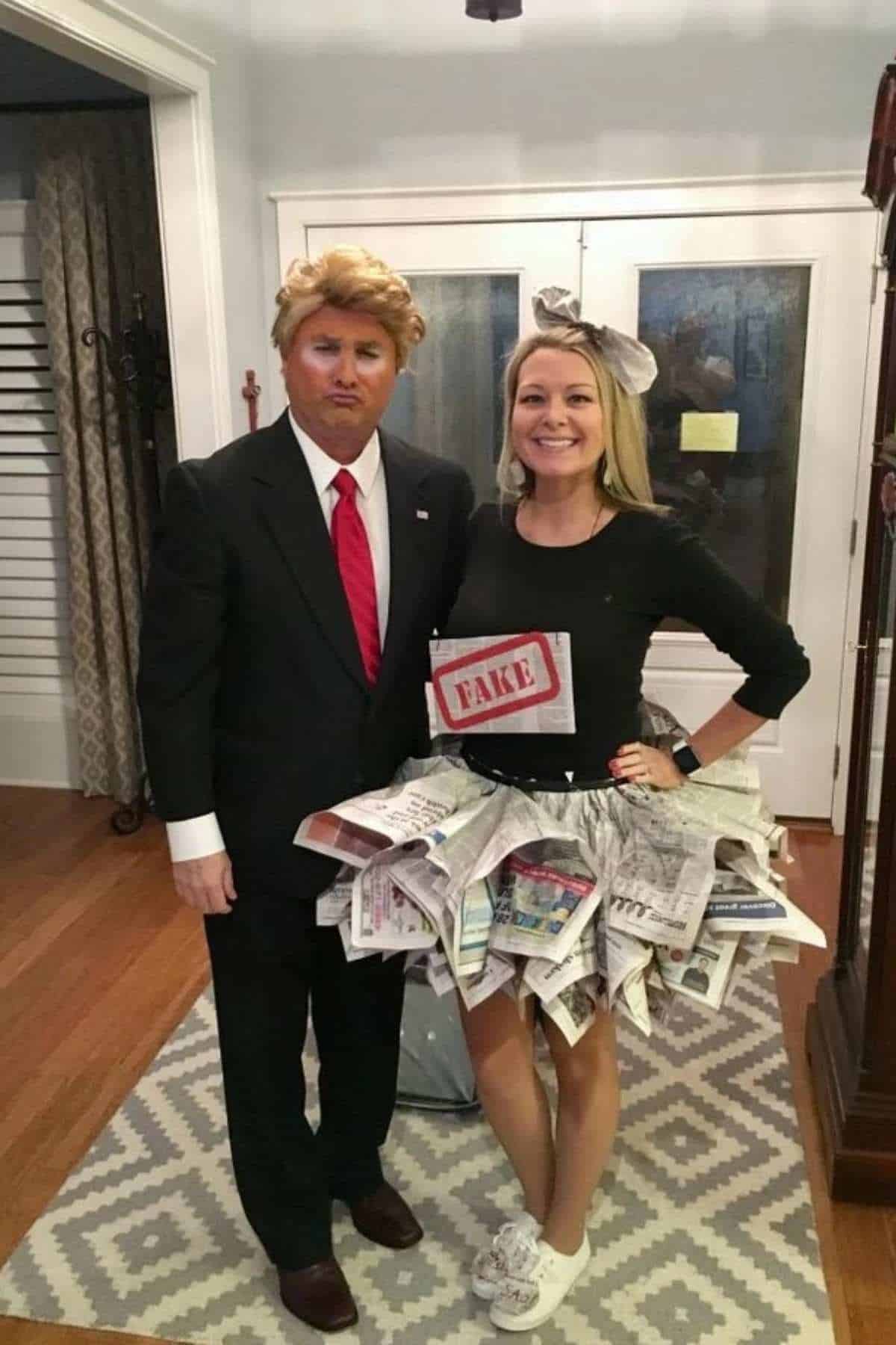 Two people in costumes: one dressed as a man in a suit with a blond wig, the other wearing a skirt made of newspapers and a "FAKE" sign, standing indoors.