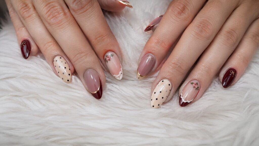 Hands with almond-shaped nails featuring burgundy, beige, and nude polish, gold tips, polka dots, and small floral designs, resting on a white fluffy surface.