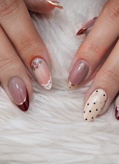 Hands with almond-shaped nails featuring burgundy, beige, and nude polish, gold tips, polka dots, and small floral designs, resting on a white fluffy surface.