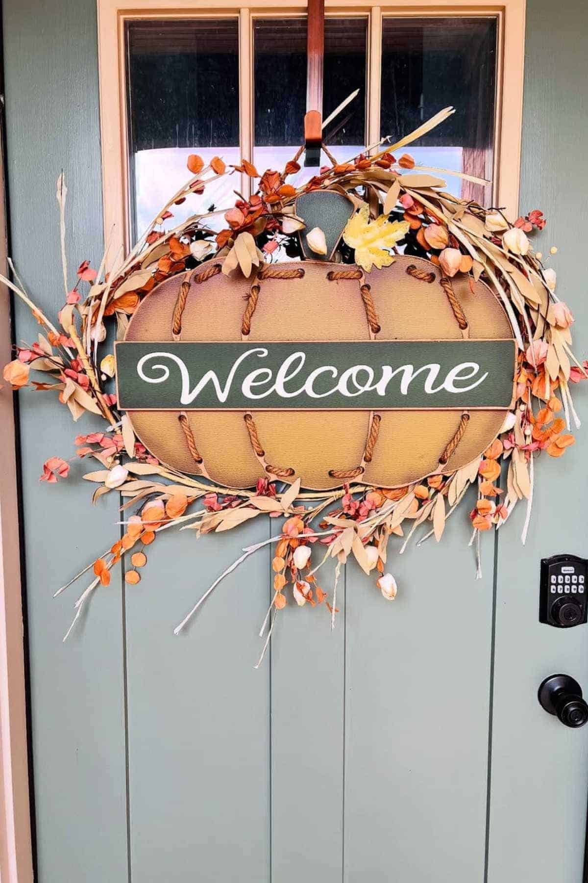 A pumpkin-shaped wreath with orange and yellow accents and a "Welcome" sign hangs on a green door with a keypad lock.