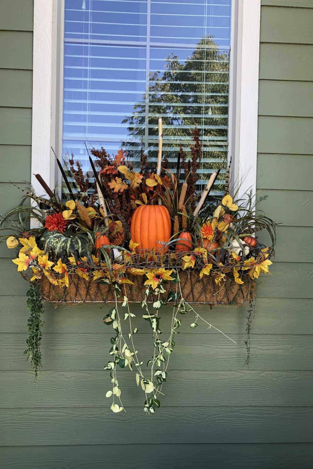 A window box filled with artificial autumn leaves, flowers, and a central orange pumpkin, beneath a window with blinds on a green exterior wall.
