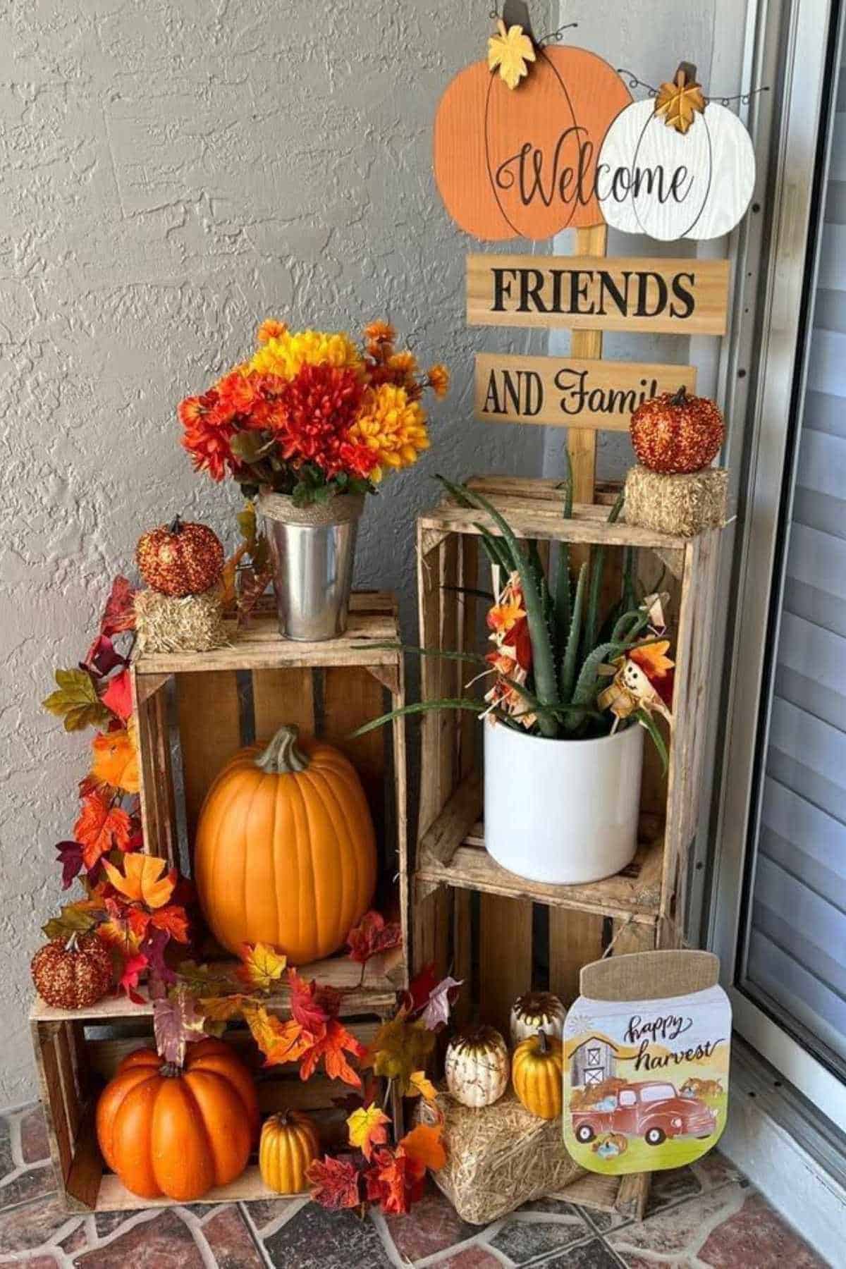 Autumn-themed porch display with pumpkins, artificial flowers, potted plants, and wooden signs reading "Welcome Friends and Family" and "Happy Harvest" arranged on wooden crates.