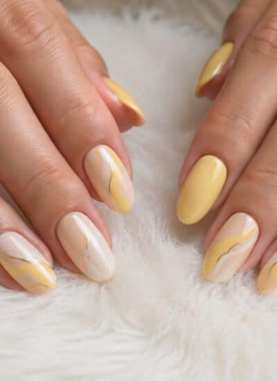 Two hands with almond-shaped nails featuring yellow and white polish with gold accent lines, resting on a white, fluffy surface.