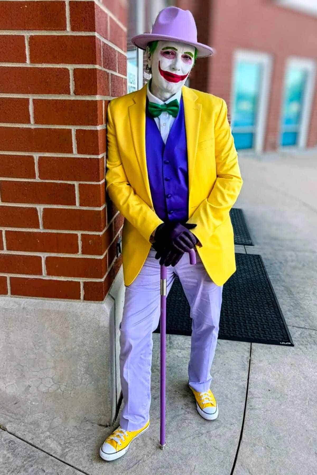Person in Joker-inspired costume with yellow jacket, purple vest, lavender pants, green bow tie, and hat, leaning against a brick wall, holding a cane.