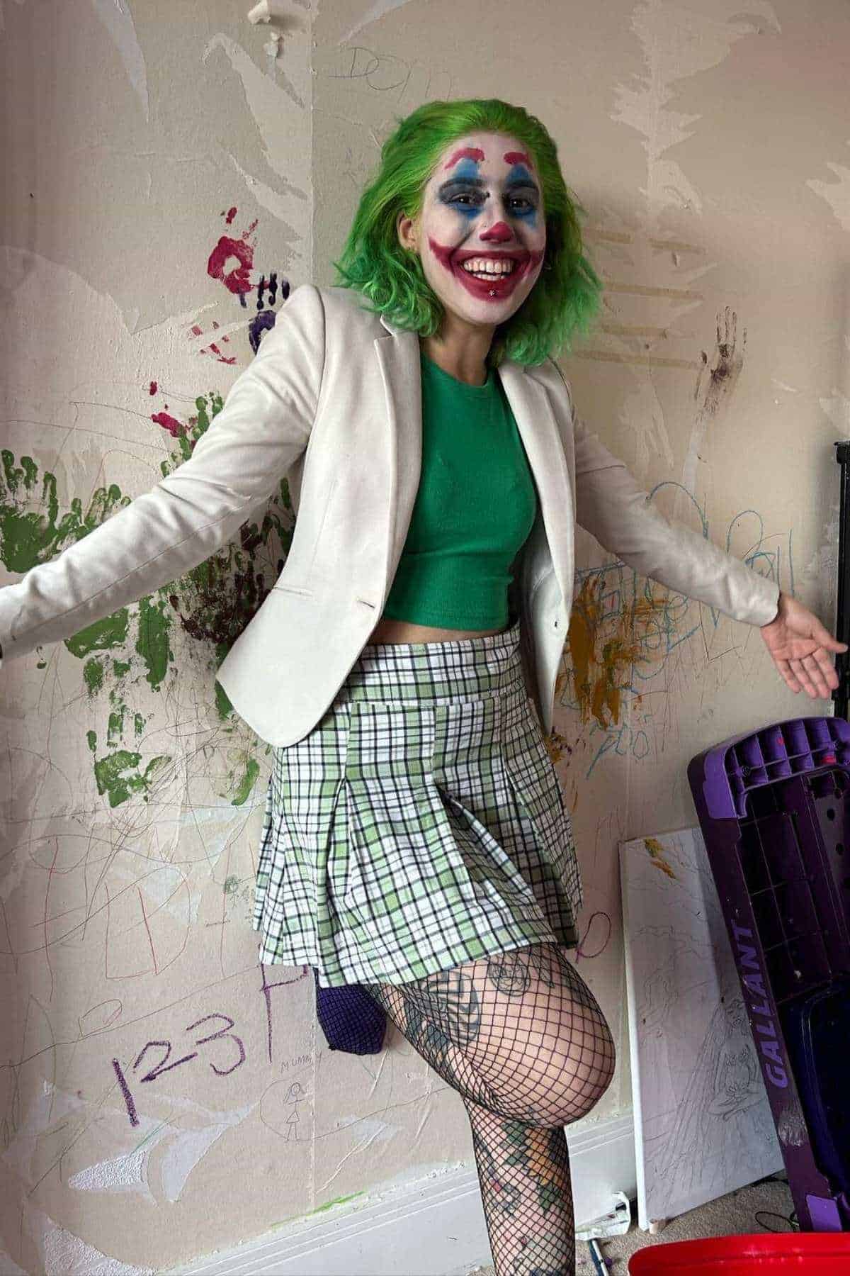 Person with green hair and clown makeup stands in front of a wall covered in colorful doodles, wearing a white blazer, green top, plaid skirt, and fishnet stockings, arms outstretched.