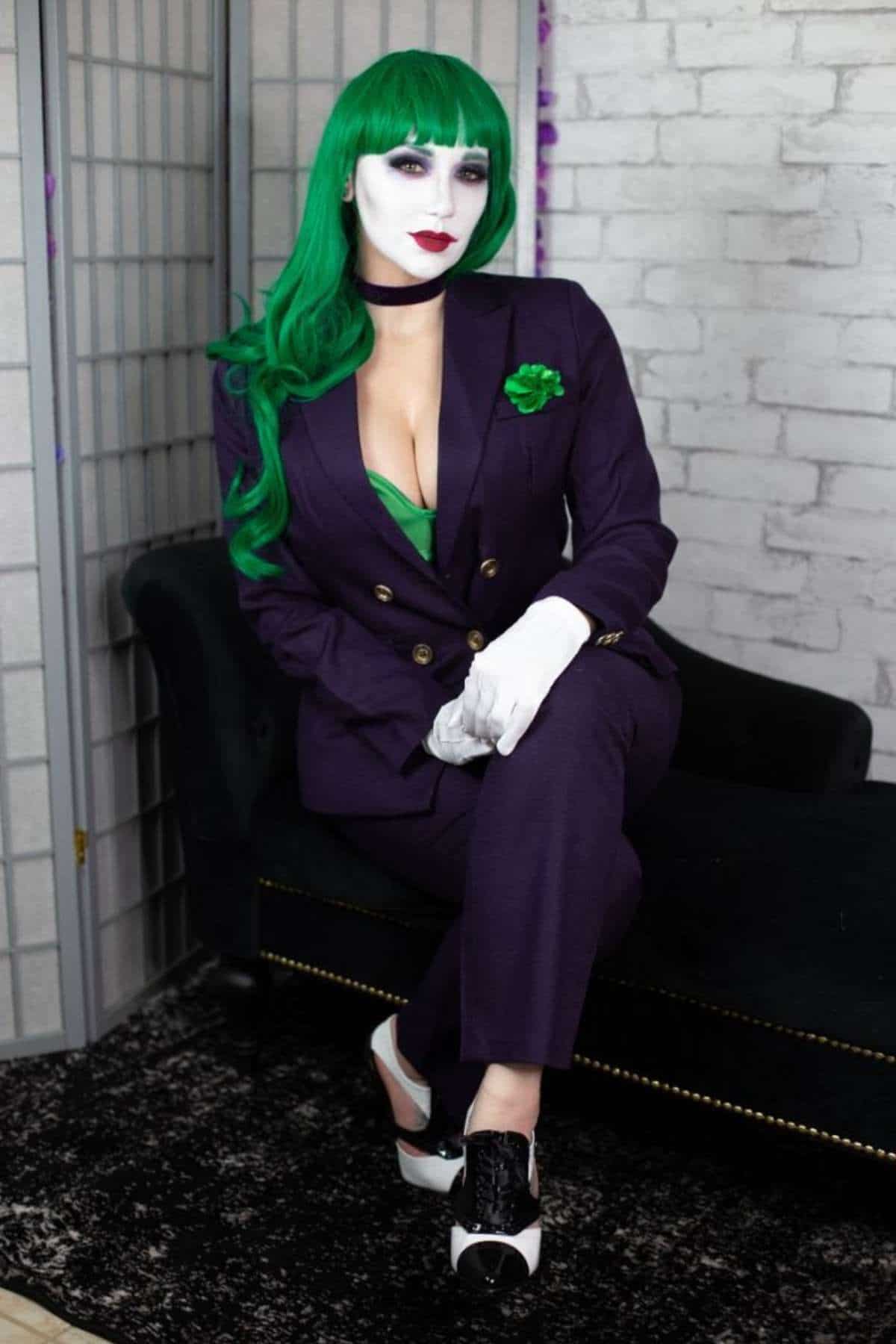 Person in a purple suit with green wig, white face makeup, and black heels sits on a dark couch against a light brick wall, resembling a comic book villain.