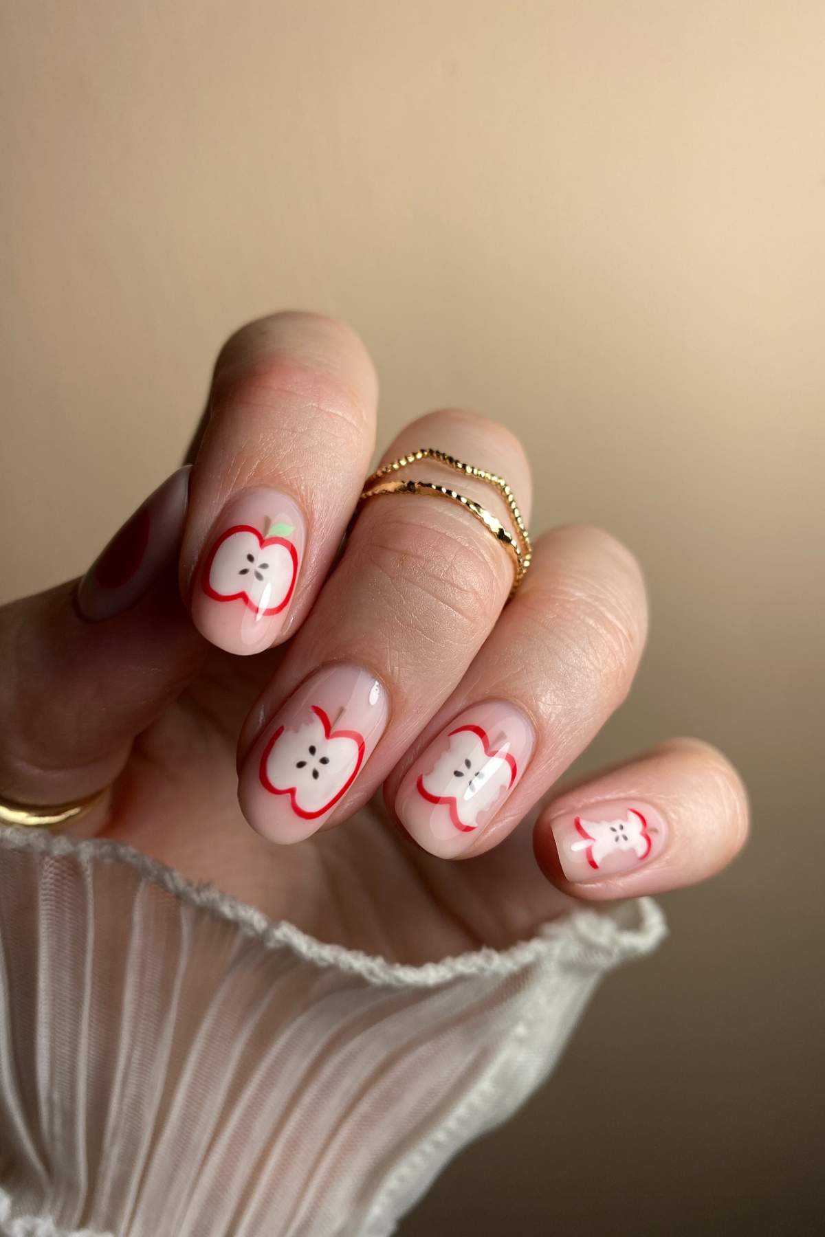 A hand with gold rings shows fingernails painted with red and white apple slice designs on a neutral background.