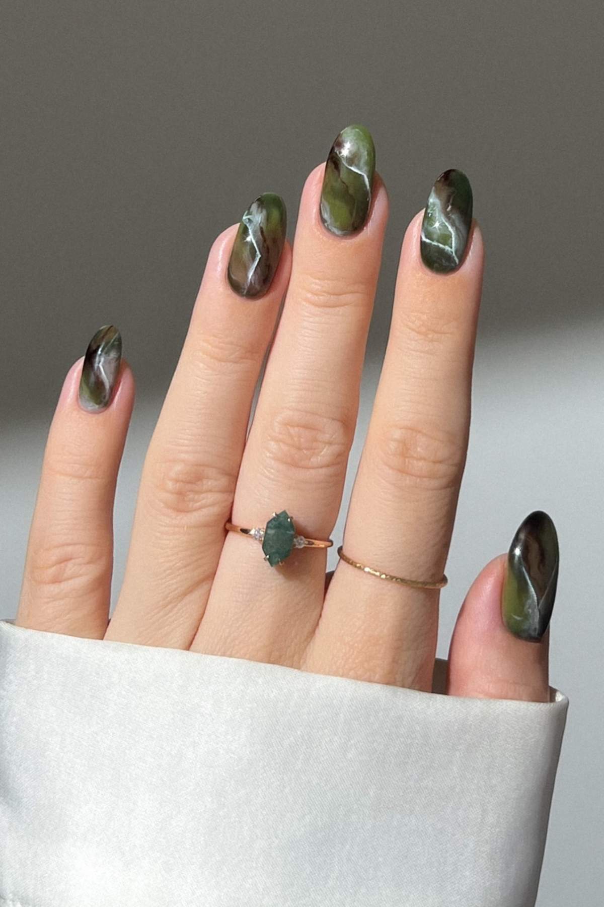 A hand with almond-shaped nails featuring a green marble design. The person wears two delicate gold rings, one with a green stone, and a white shirt sleeve partially covers the wrist.