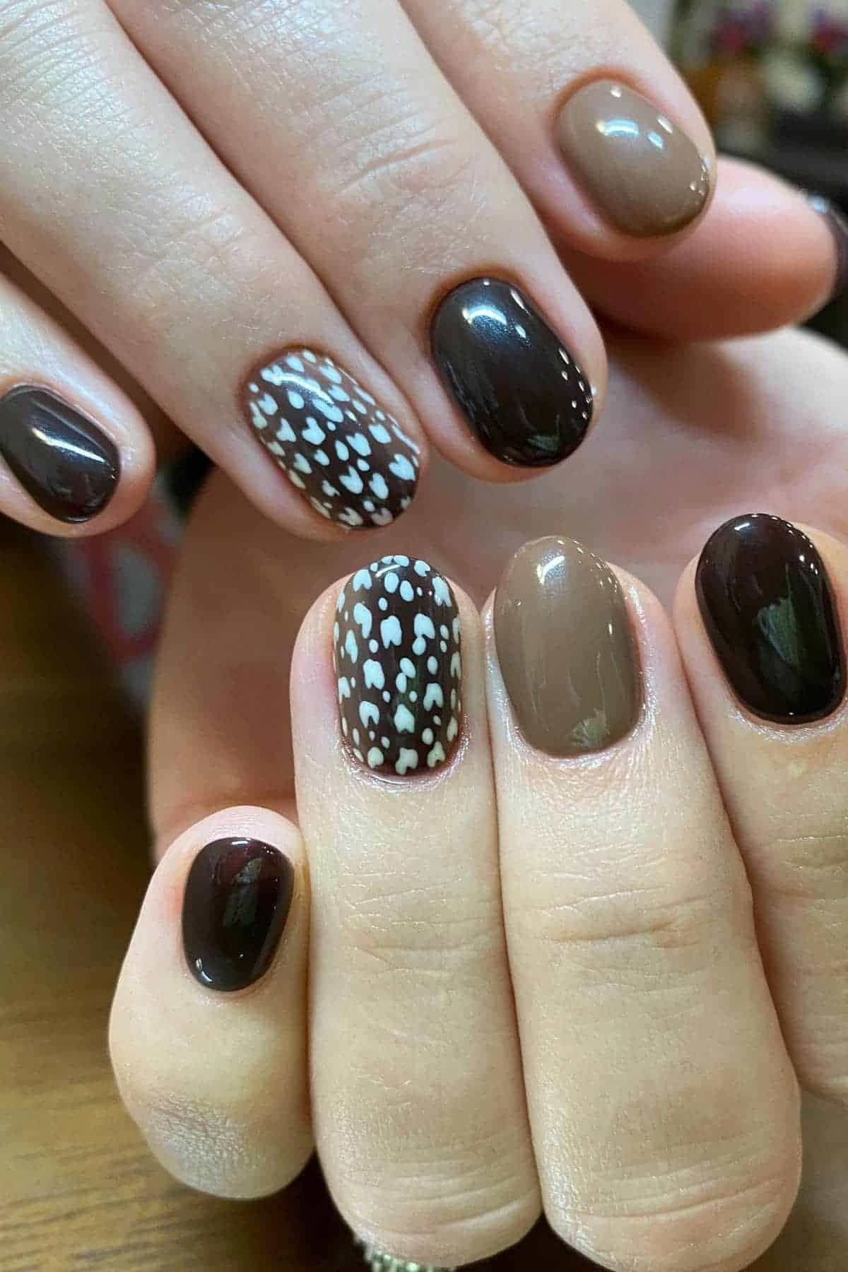 Close-up of hands with short, oval nails painted in brown, beige, and dark brown shades, featuring white polka dot and heart patterns on two accent nails.