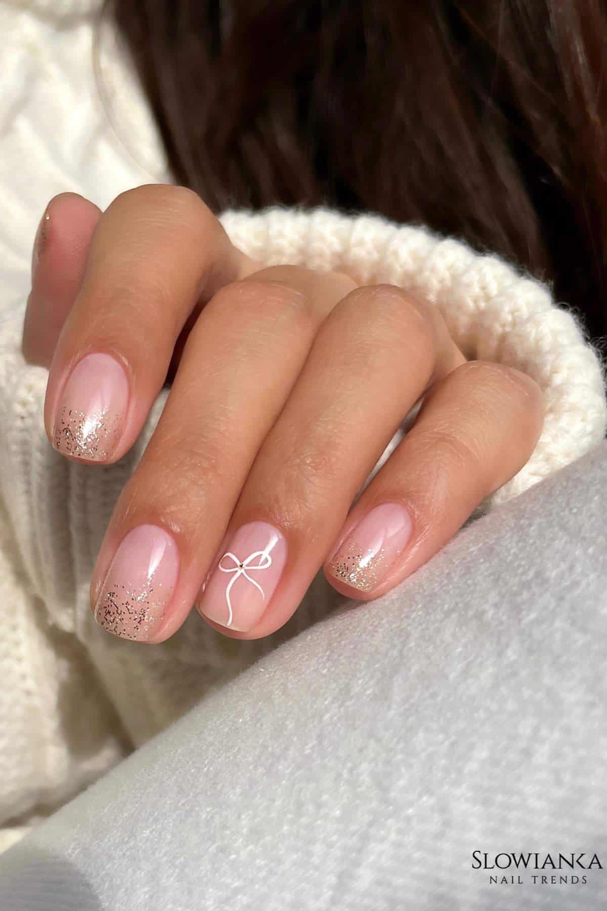 A hand with manicured nails features a light pink polish, glitter tips, and a white bow design on the ring finger.