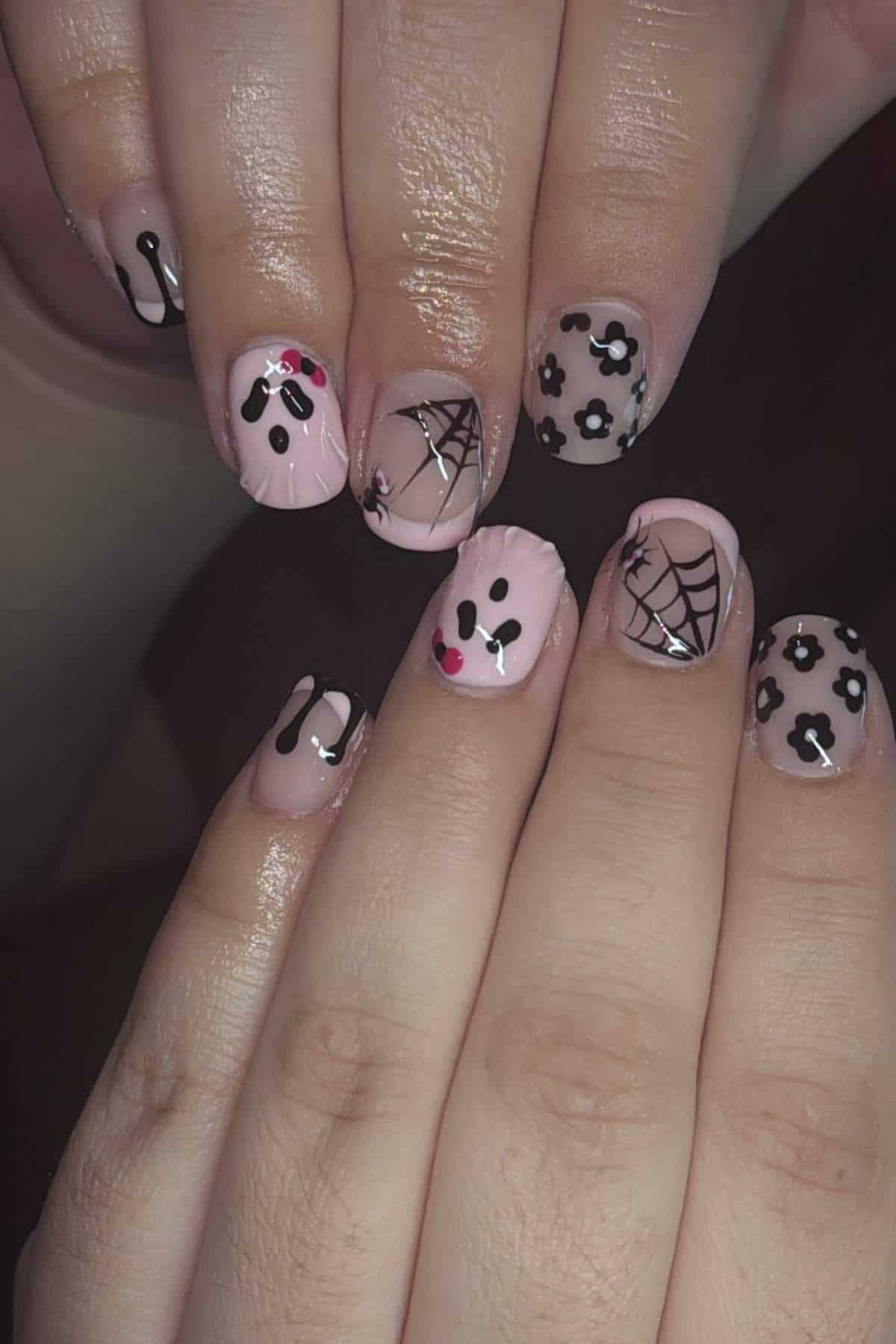 Hands with pink nail polish featuring black cow spots, spider webs, drips, and cartoon ghost designs on each fingernail.