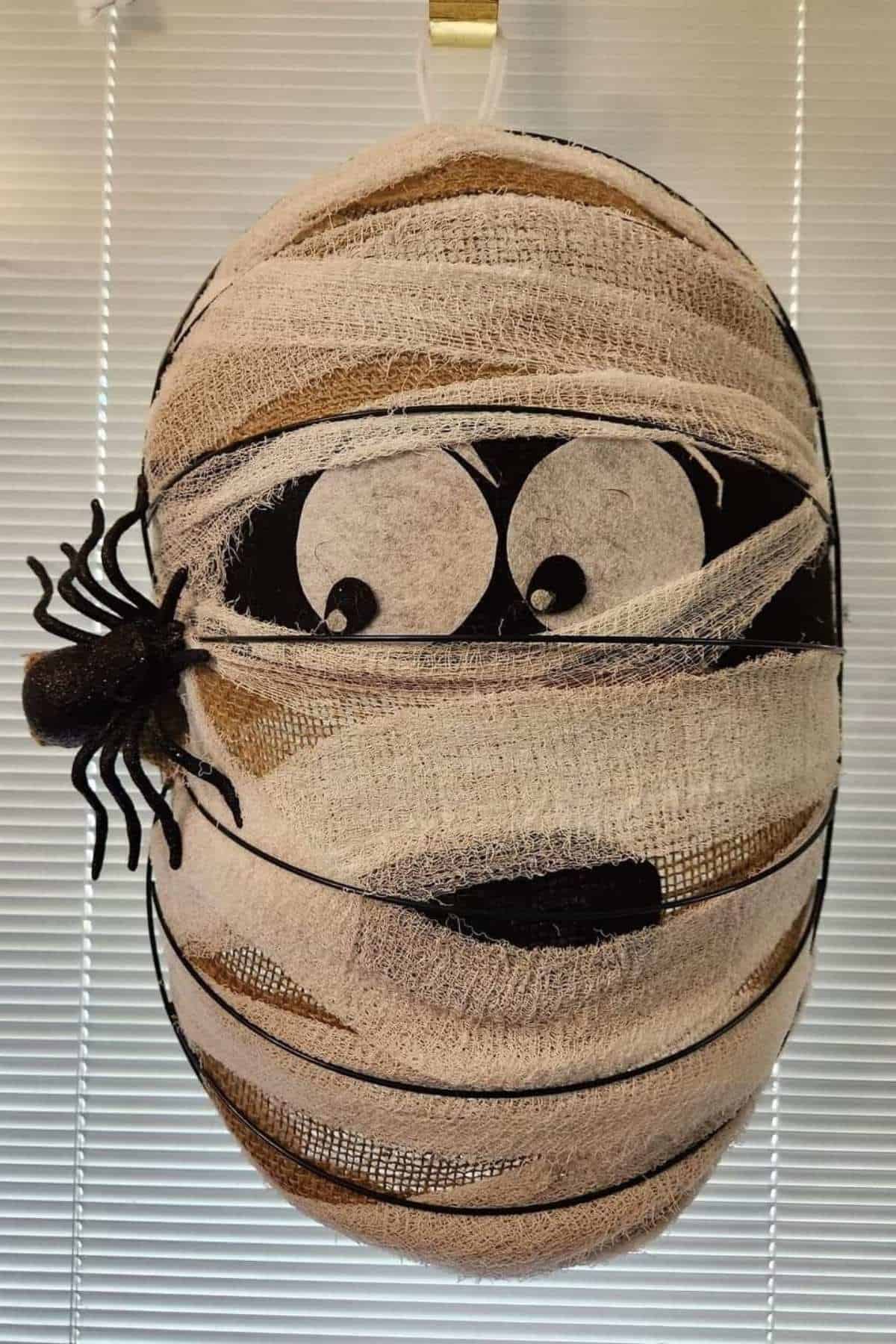 A mummy face decoration with googly eyes, bandages, and a small black spider on the left side, hanging in front of window blinds.