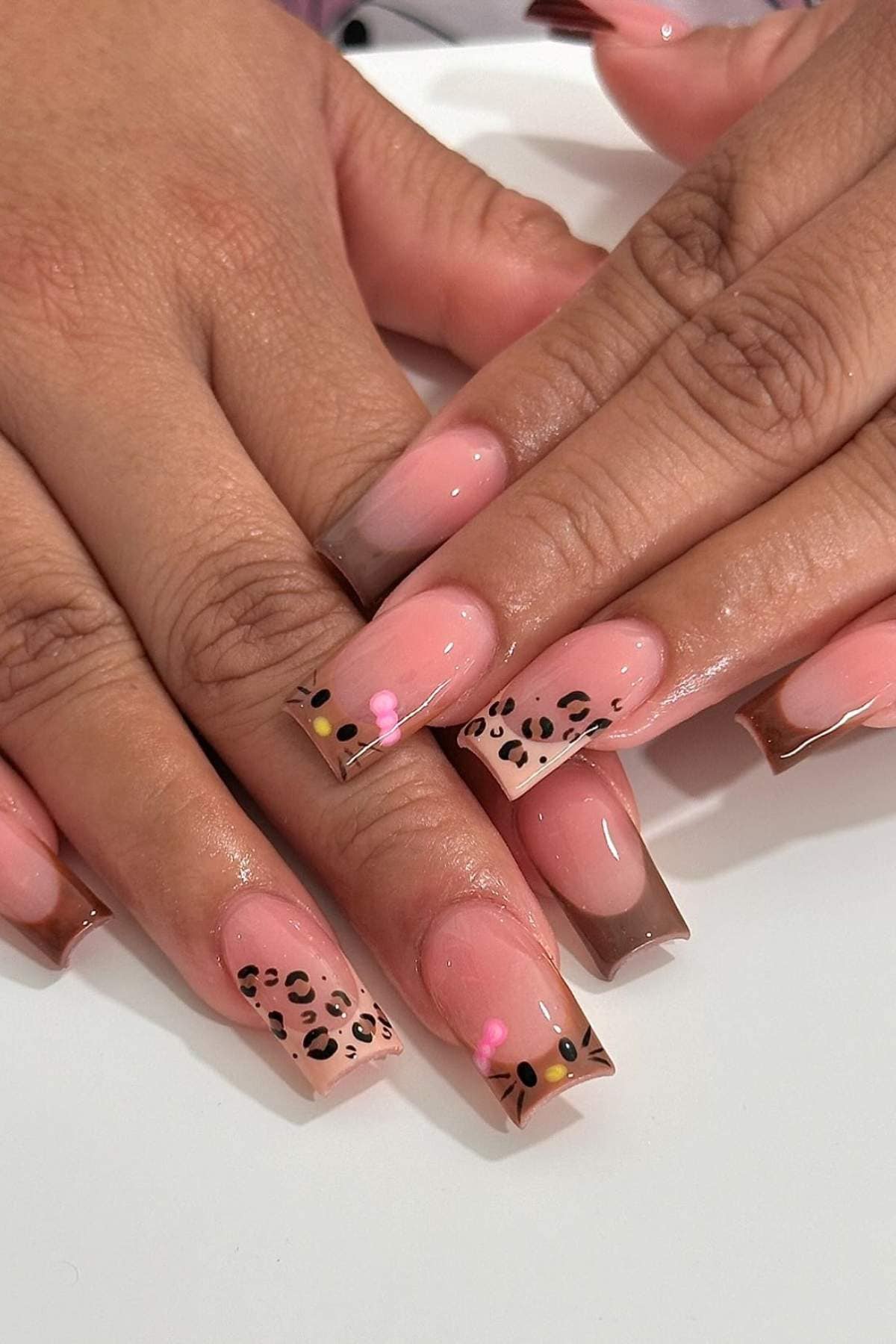 Hands with square-shaped acrylic nails. Nails have a nude base with brown French tips, some featuring leopard print accents and small pink dots on a white surface.