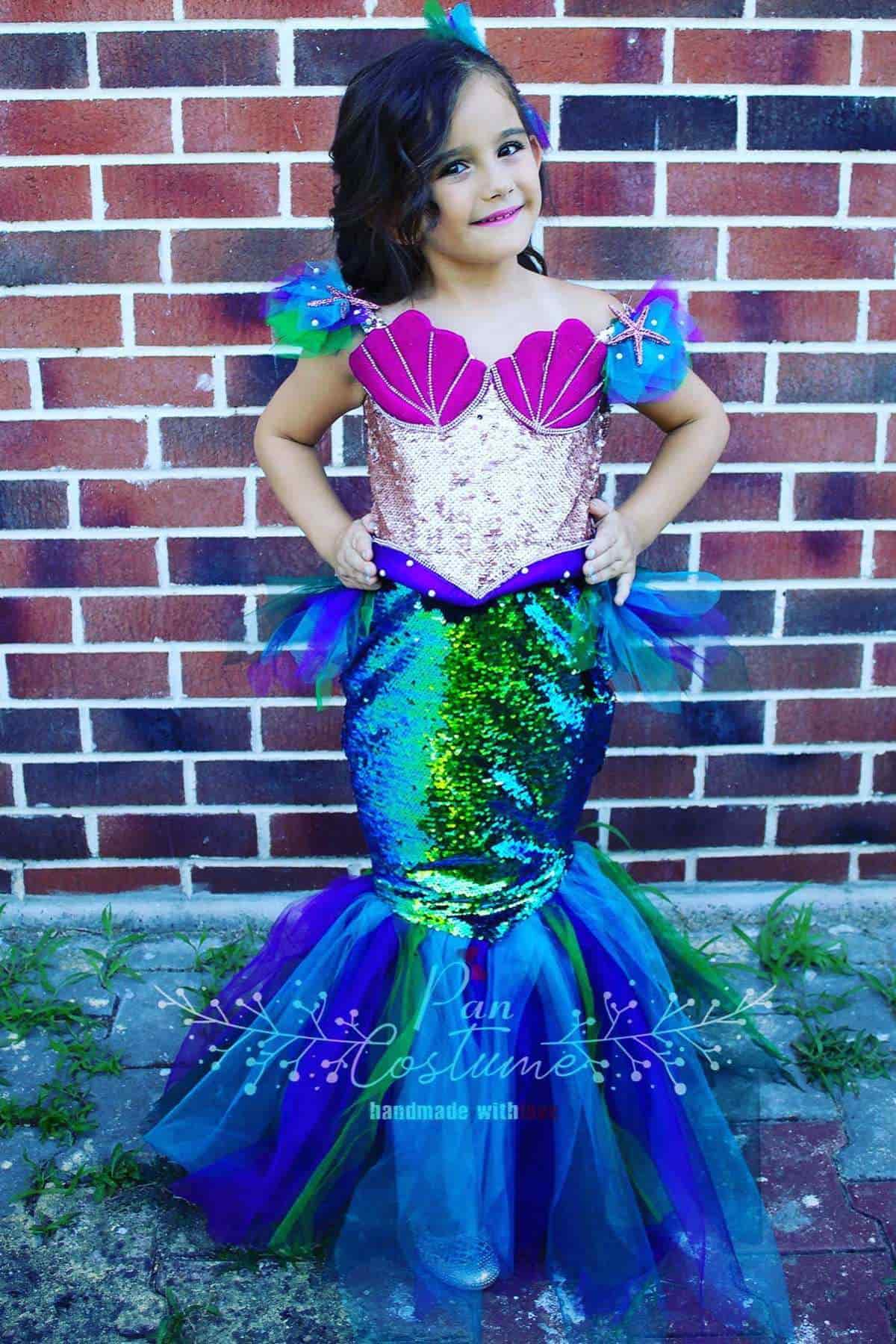 Young child in a colorful mermaid costume with a sequined tail and a shell-patterned top stands in front of a brick wall.