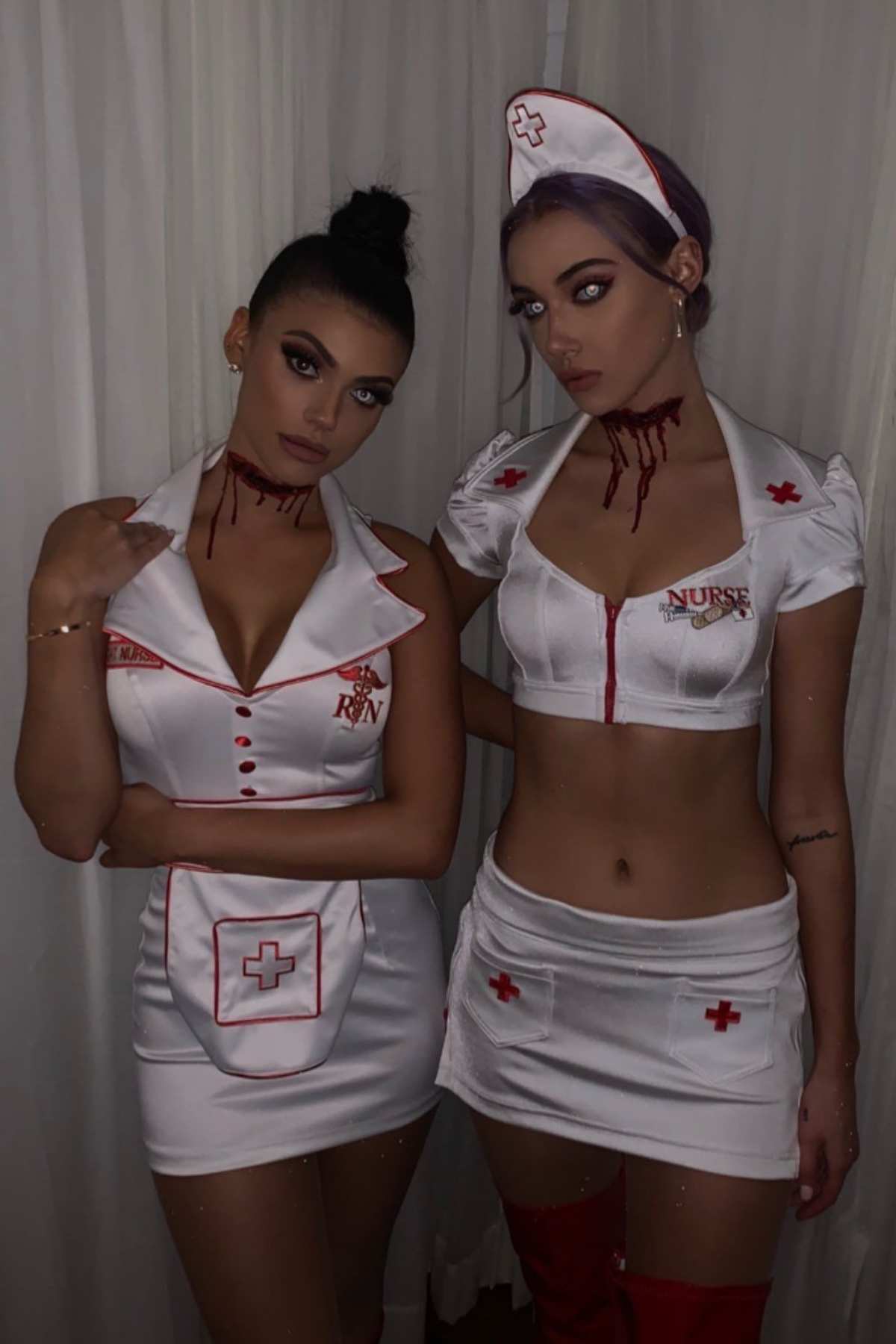 Two women in white nurse costumes with red accents and fake blood on their necks stand side by side in front of a white curtain.