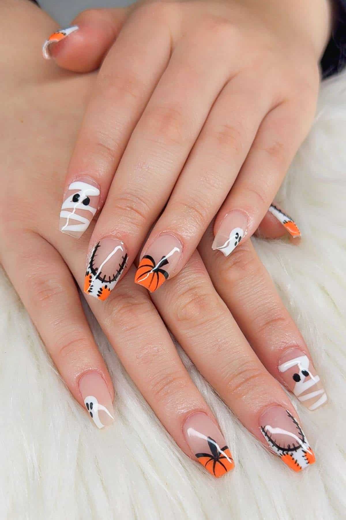 Hands with long nails featuring Halloween-themed nail art, including mummies, ghosts, spiders, pumpkins, and stitched patterns in black, white, and orange on a white fur surface.