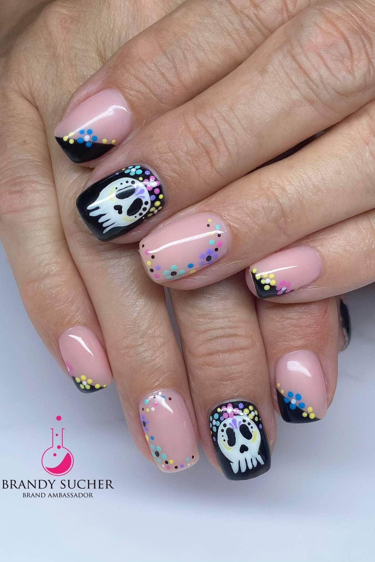 Fingernails with black French tips, multicolored dot accents, and skull designs on two nails. The skin tone is fair and the nails are almond-shaped.