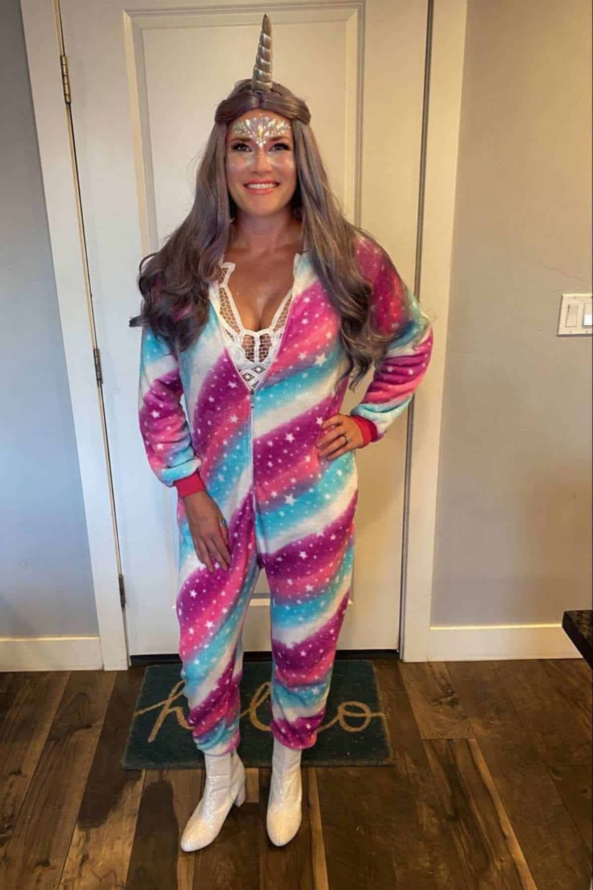 Person standing indoors wearing a colorful unicorn onesie, unicorn horn headband, white boots, and glittery face paint, with long silver hair and hands by their sides.