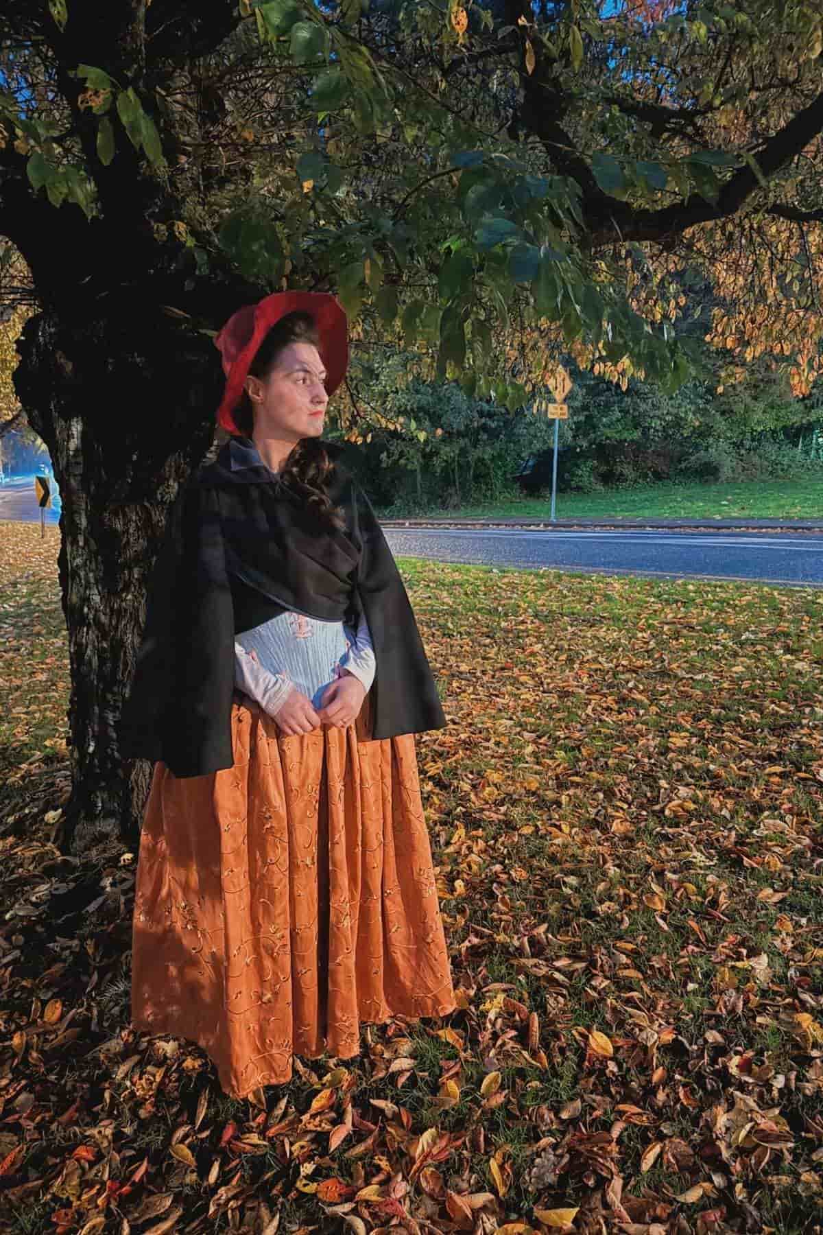 A woman in historical clothing stands under a tree with autumn leaves on the ground, looking into the distance near a road.