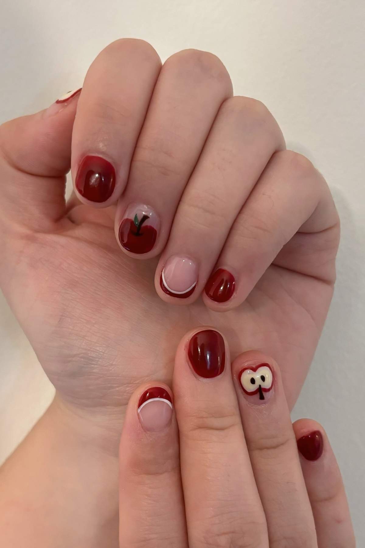 Hands with red nail polish; some nails feature apple-themed designs, including a whole apple with a stem, a half apple with seeds, and red French tips.