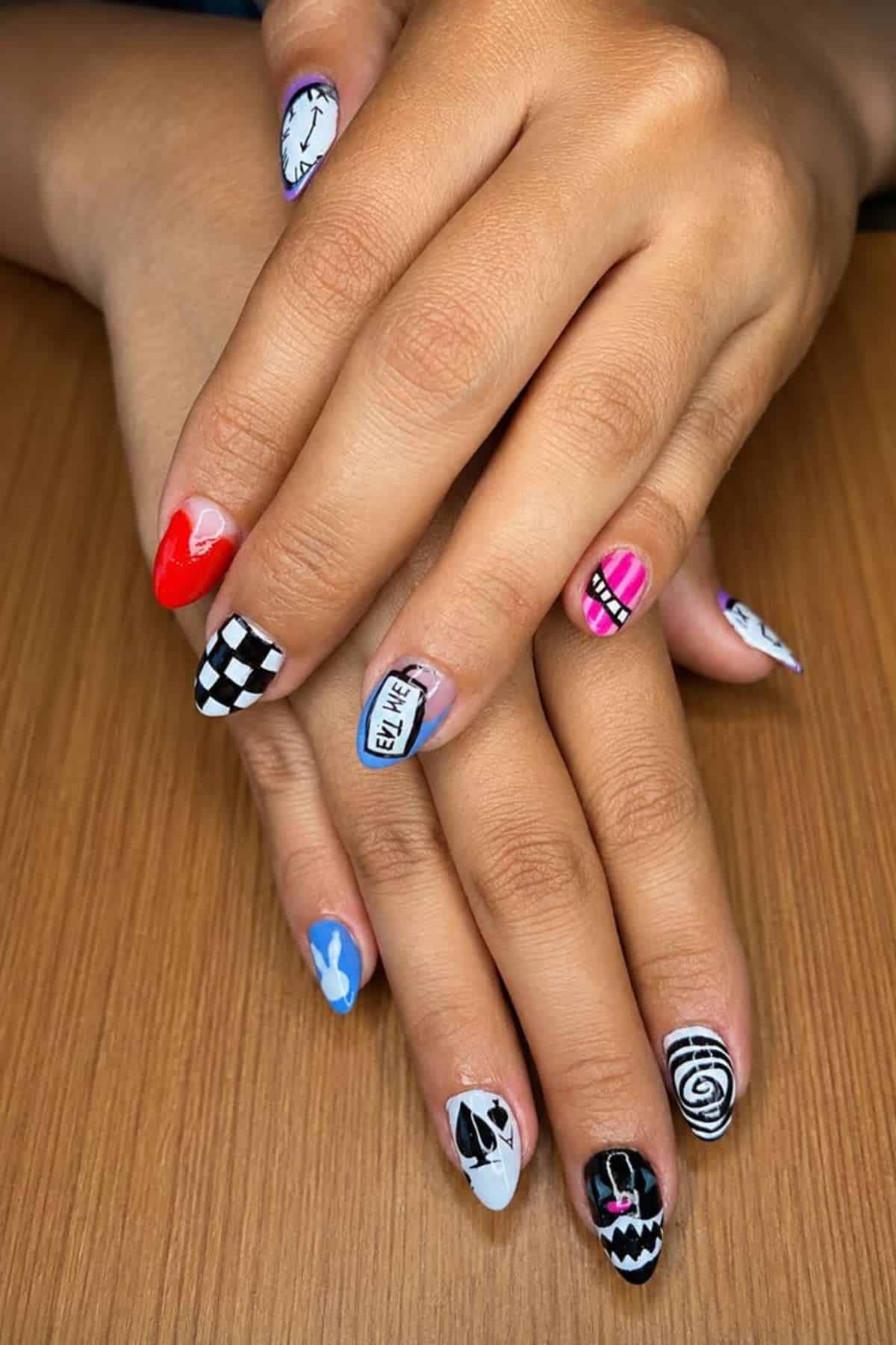Hands with almond-shaped nails featuring various colorful and patterned nail art designs, including hearts, stripes, checkerboard, and cartoon-like elements, resting on a wooden surface.