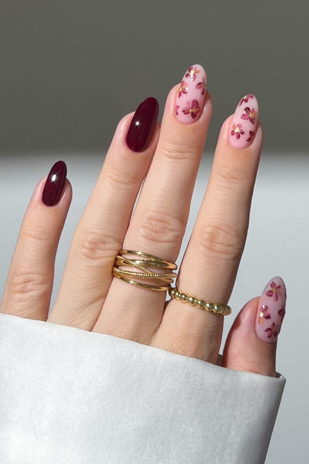 A hand with gold rings displays manicured nails; three nails have pink polish with small flower designs, and two nails are painted dark burgundy.