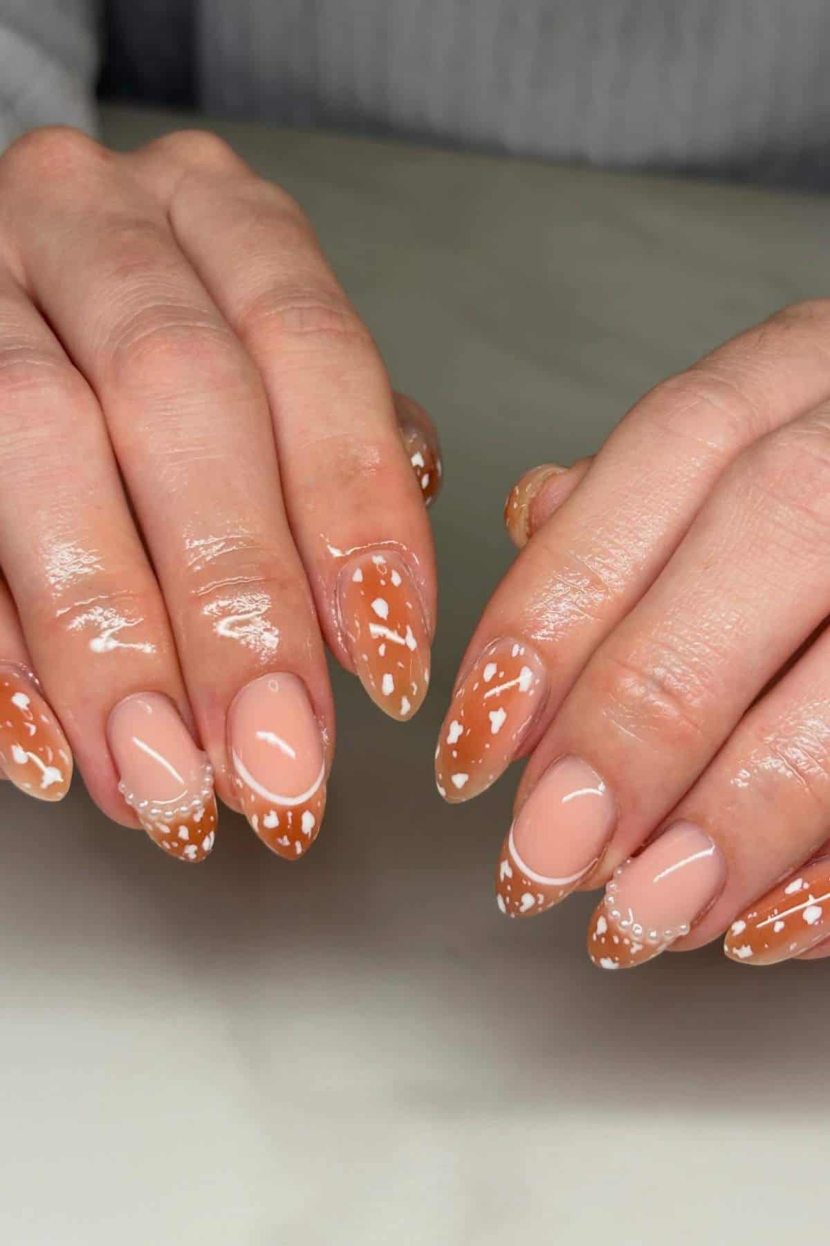 Two hands with almond-shaped nails, featuring a glossy nude base and orange tips with small white floral rhinestone designs, resting on a marble surface.