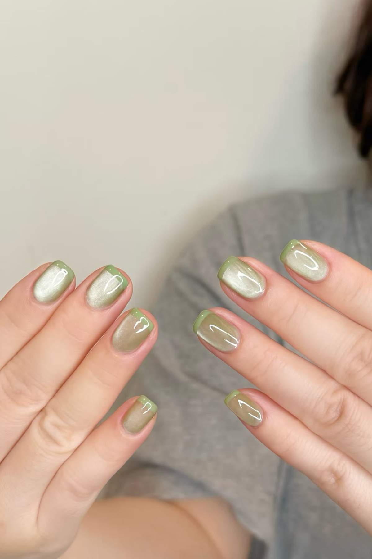 Close-up of hands displaying short, oval-shaped nails with shimmery beige polish and thin light green tips, against a neutral background.