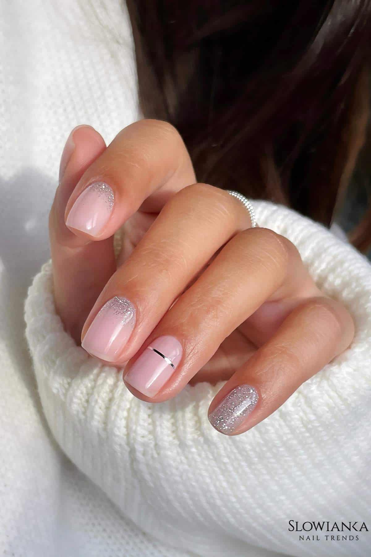 A hand with short, almond-shaped nails painted in pale pink with silver glitter accents and a thin black line on the ring finger. The person is wearing a white knitted sweater.
