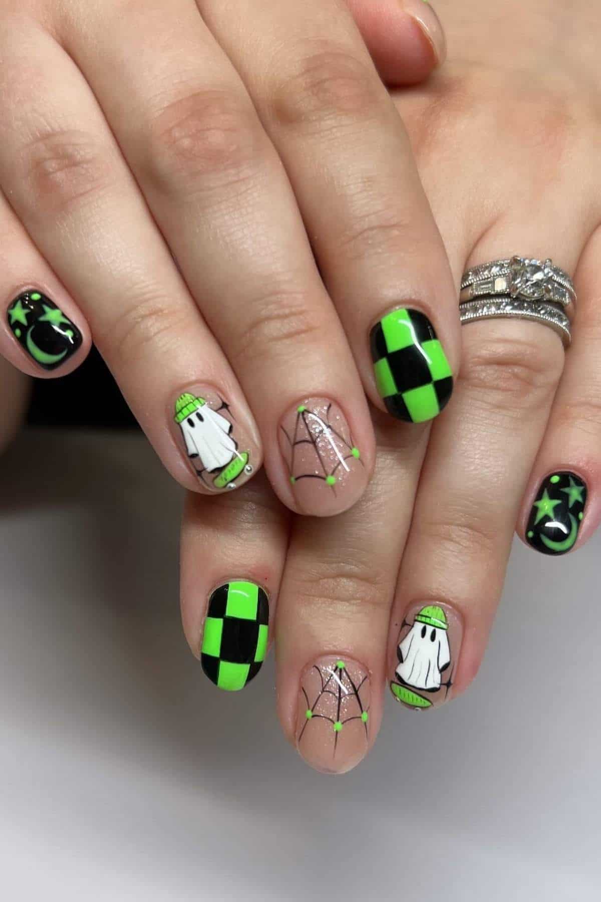 Hands with Halloween-themed nail art featuring green and black checkerboard patterns, ghosts, stars, and spiderweb designs.
