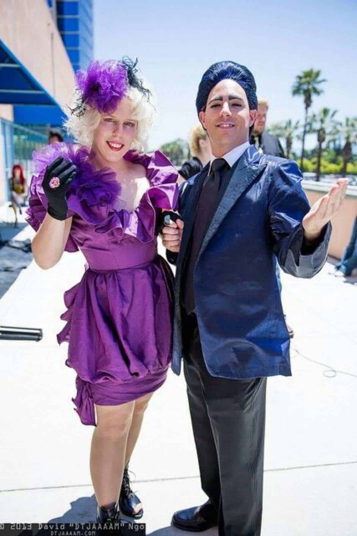 Two people in colorful, formal costumes pose outdoors; one wears a purple dress and hat, the other a blue suit and wig.