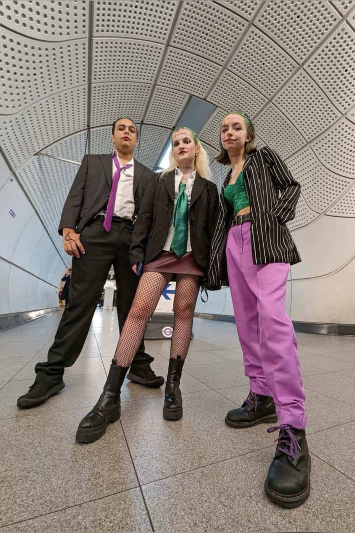 Three people pose in a tunnel wearing suits and bold outfits, including green and purple accents, fishnet tights, and chunky black boots. The photo is taken from a low angle.