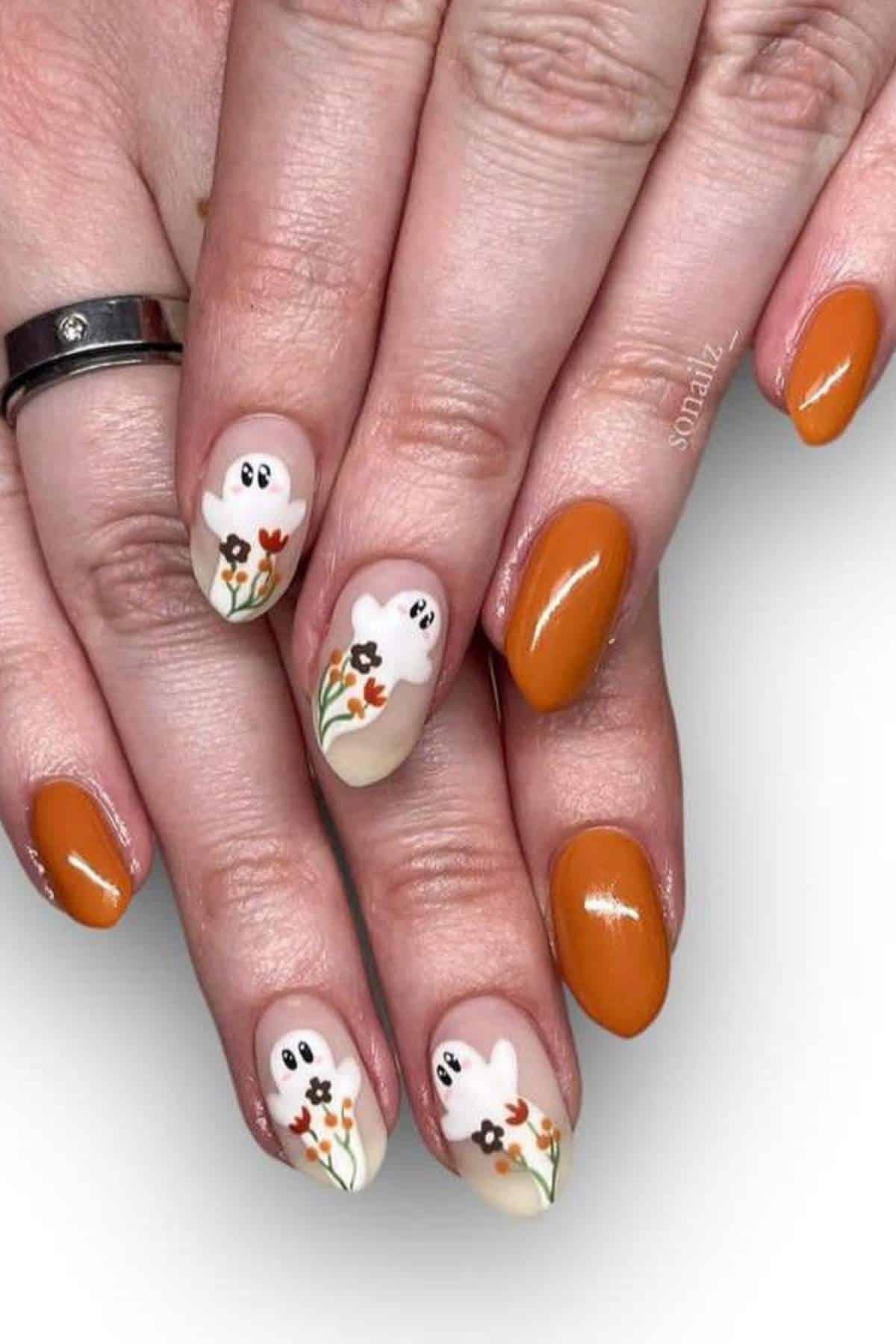Hands with burnt orange nail polish. Accent nails feature painted white ghost designs with flowers and greenery.