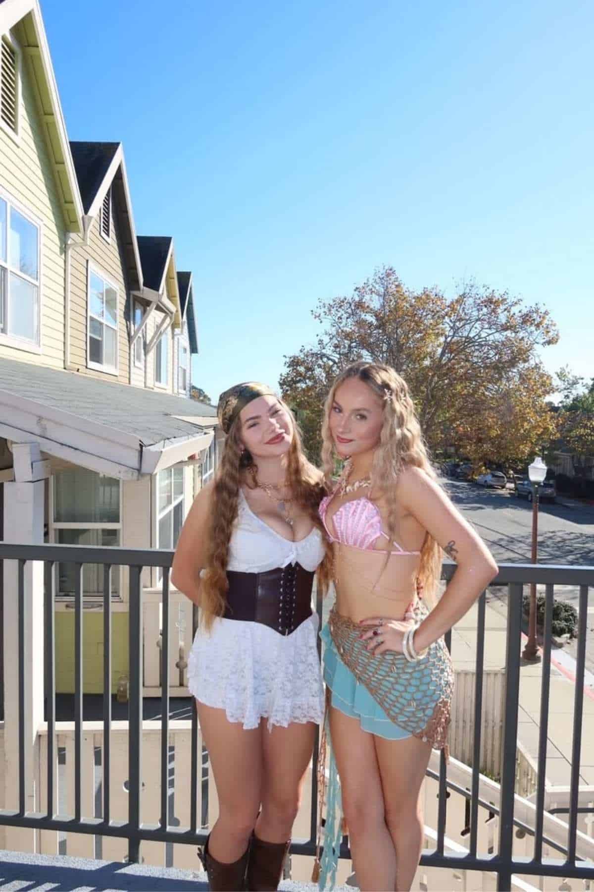 Two women in costumes pose on a sunny balcony; one wears a white lace dress with a corset, the other a shell bra top and netted skirt. Houses and trees are visible in the background.