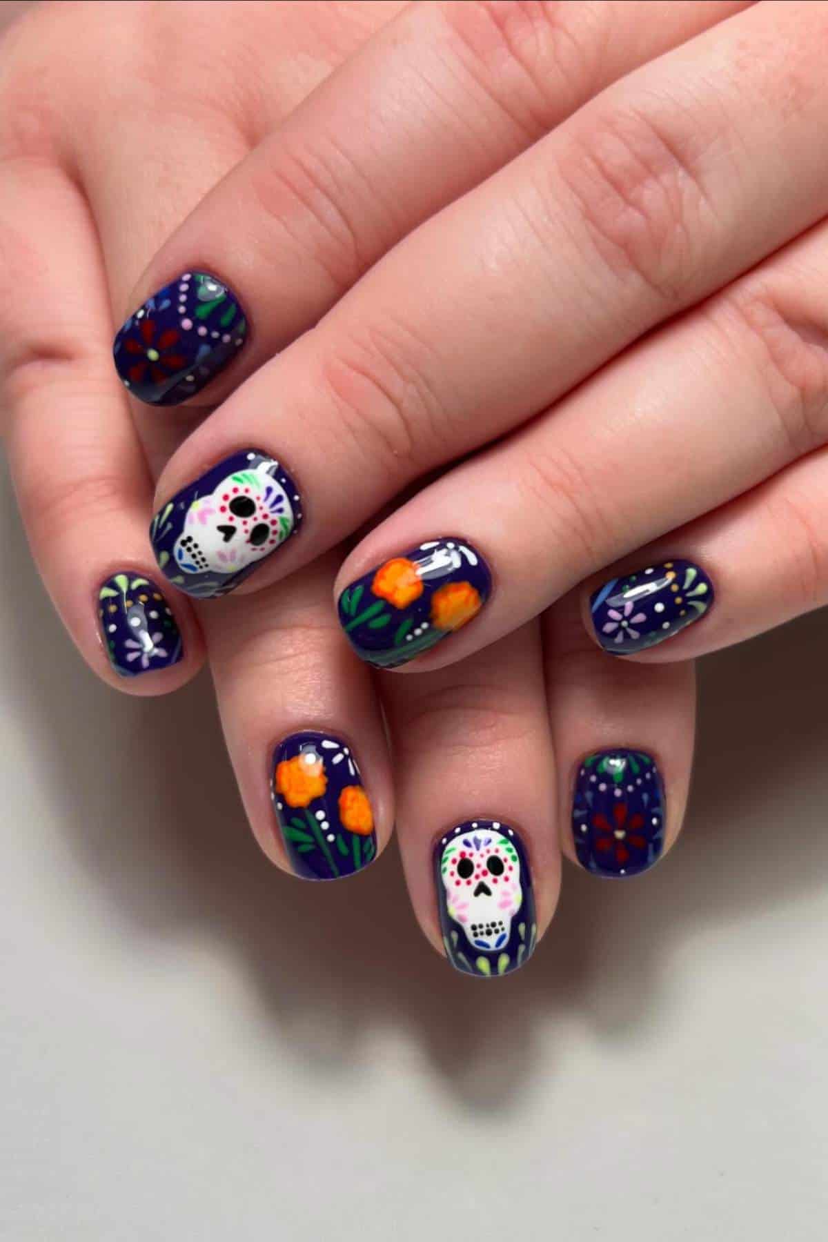 Hands with dark blue nail polish featuring Day of the Dead sugar skulls, marigolds, and floral decorative patterns on each nail.