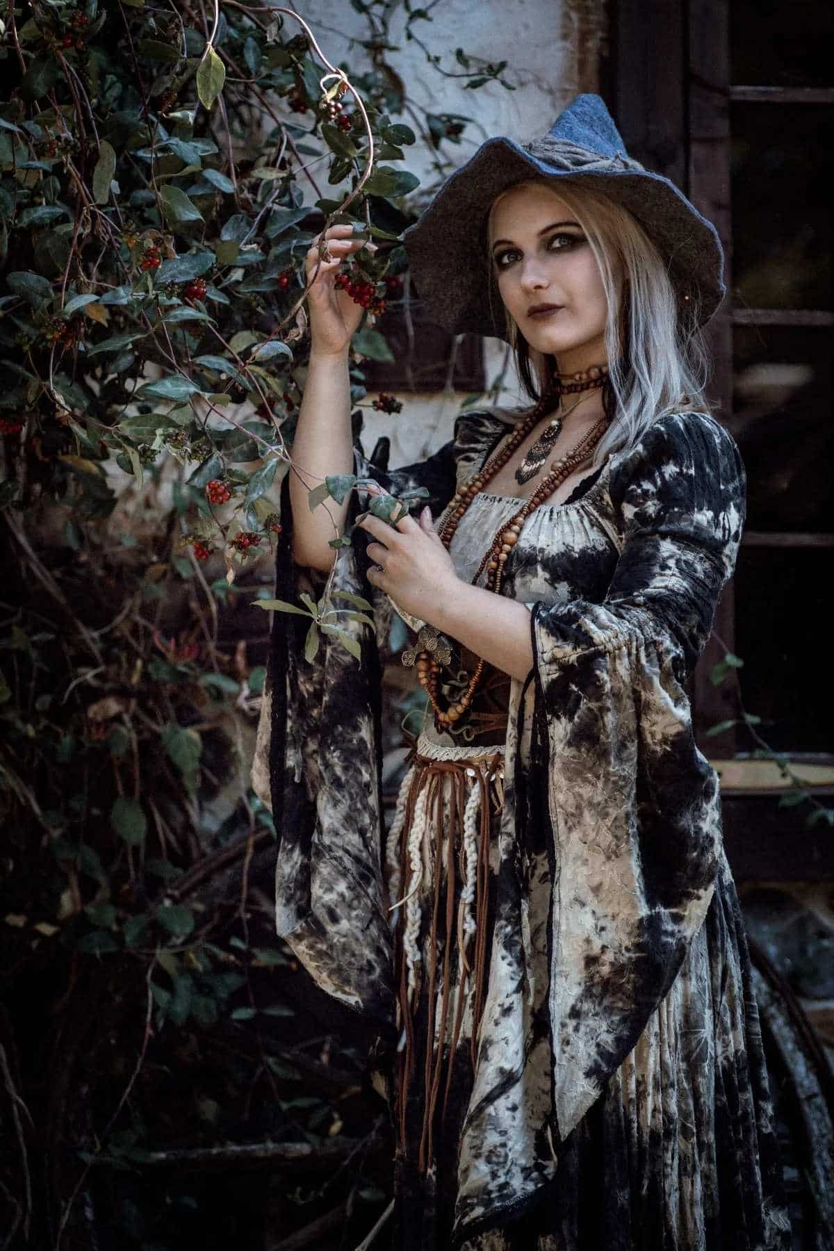 Woman in a wide-brimmed hat and velvet dress stands beside a berry bush, holding a branch and looking at the camera.