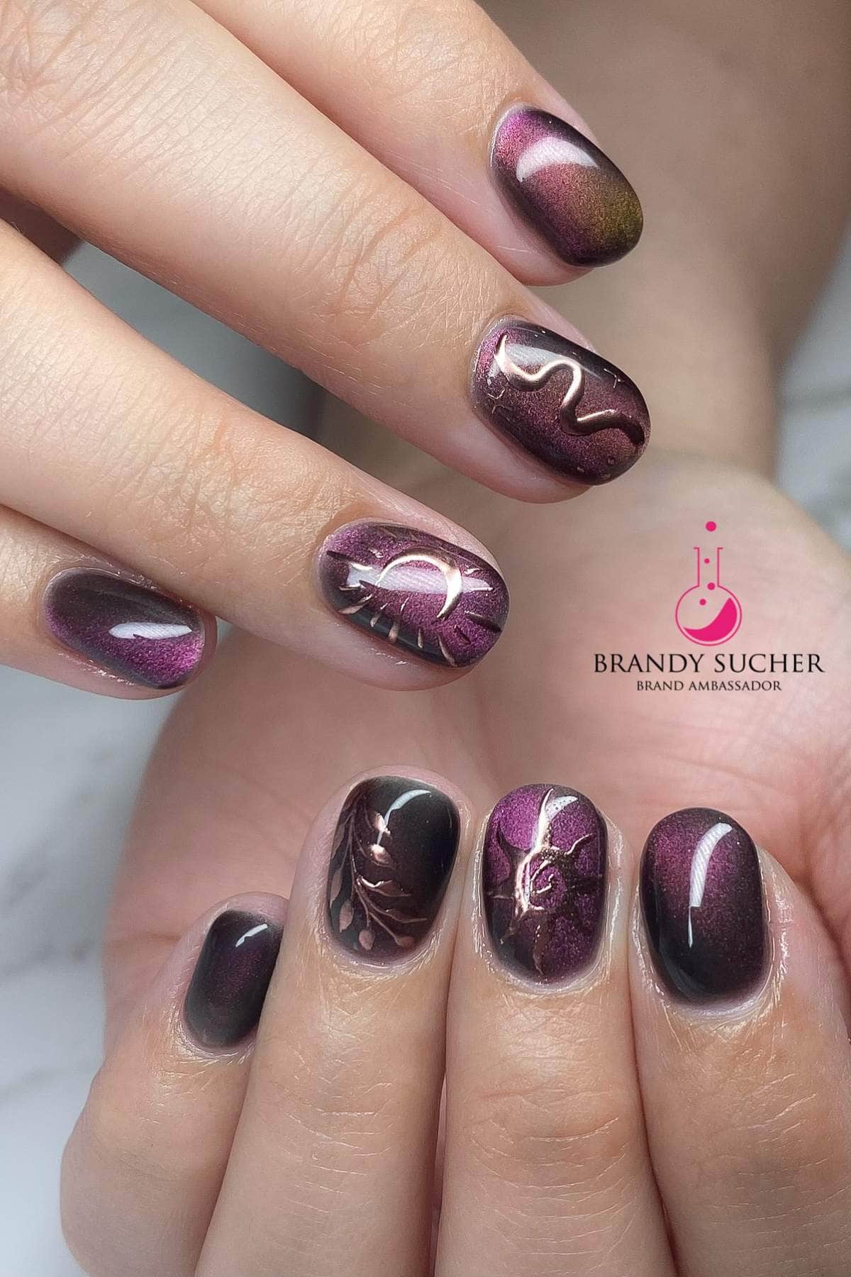 Close-up of hands with short, almond-shaped nails painted in shimmery burgundy polish, featuring metallic leaf and swirl designs, with a Brandy Sucher brand ambassador logo.