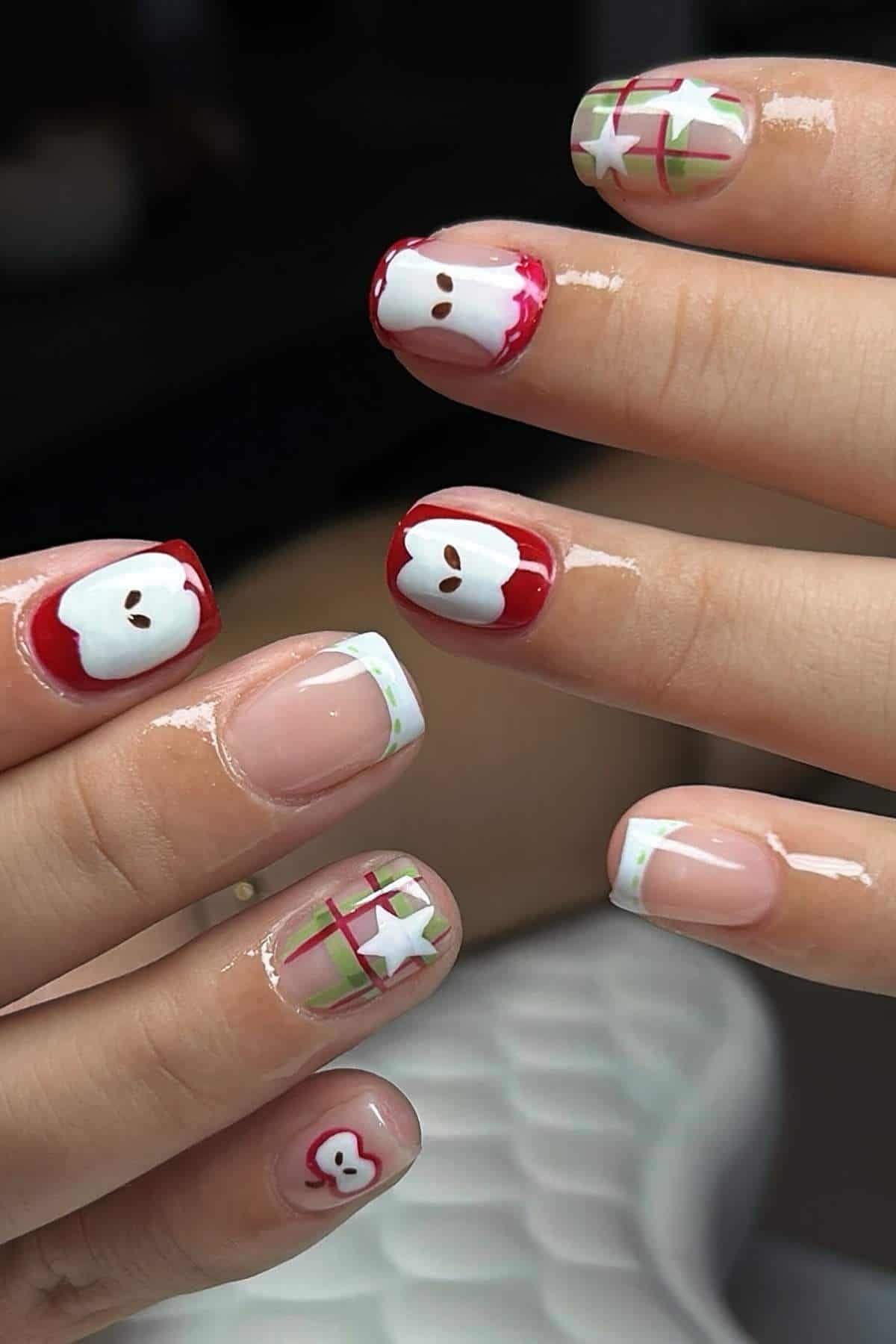 Hands with nail art; some nails feature red and white designs resembling apple slices, others have green and red plaid patterns, and one has a small white star accent.