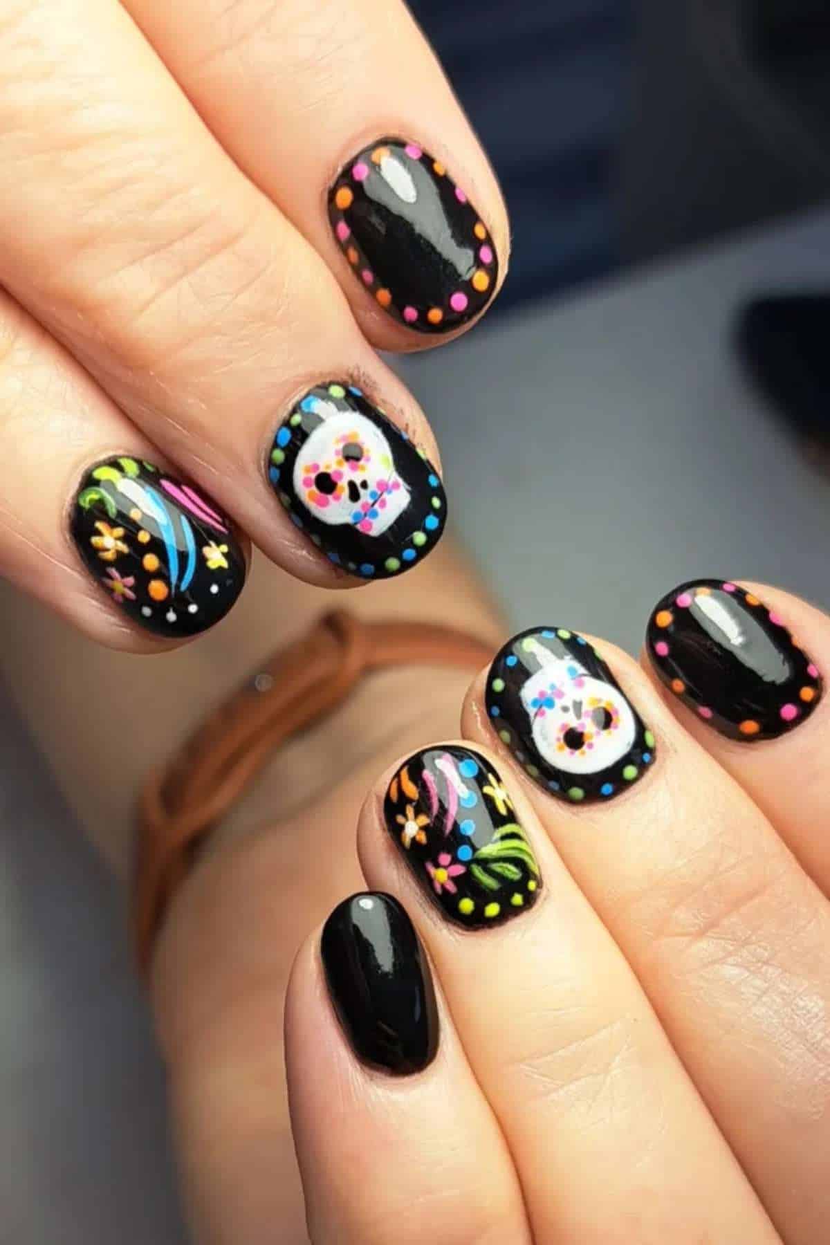 Hands with black-painted nails featuring colorful sugar skull and confetti designs, inspired by Day of the Dead, in a close-up view.
