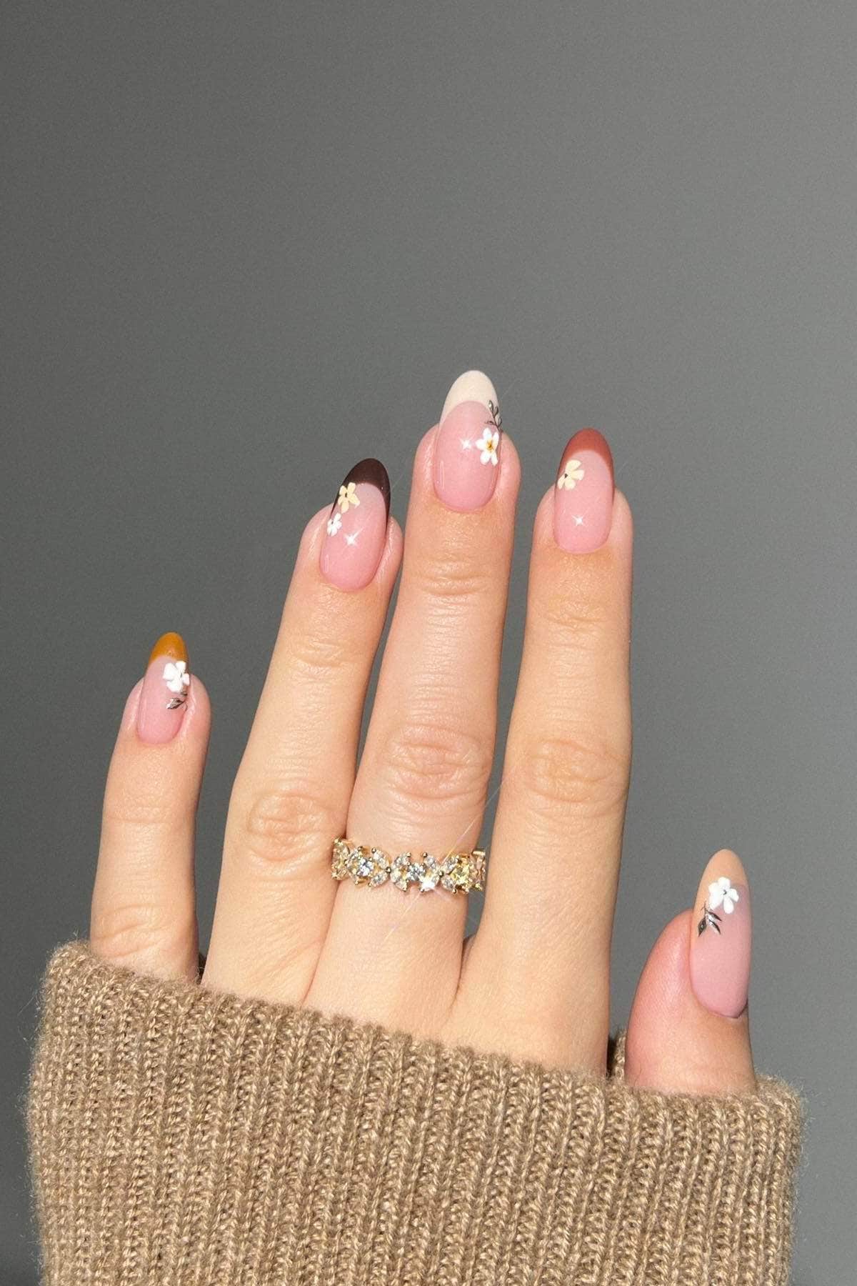 A hand with multicolored French tip nails and small flower designs wears gold rings. The person is wearing a beige knit sweater.