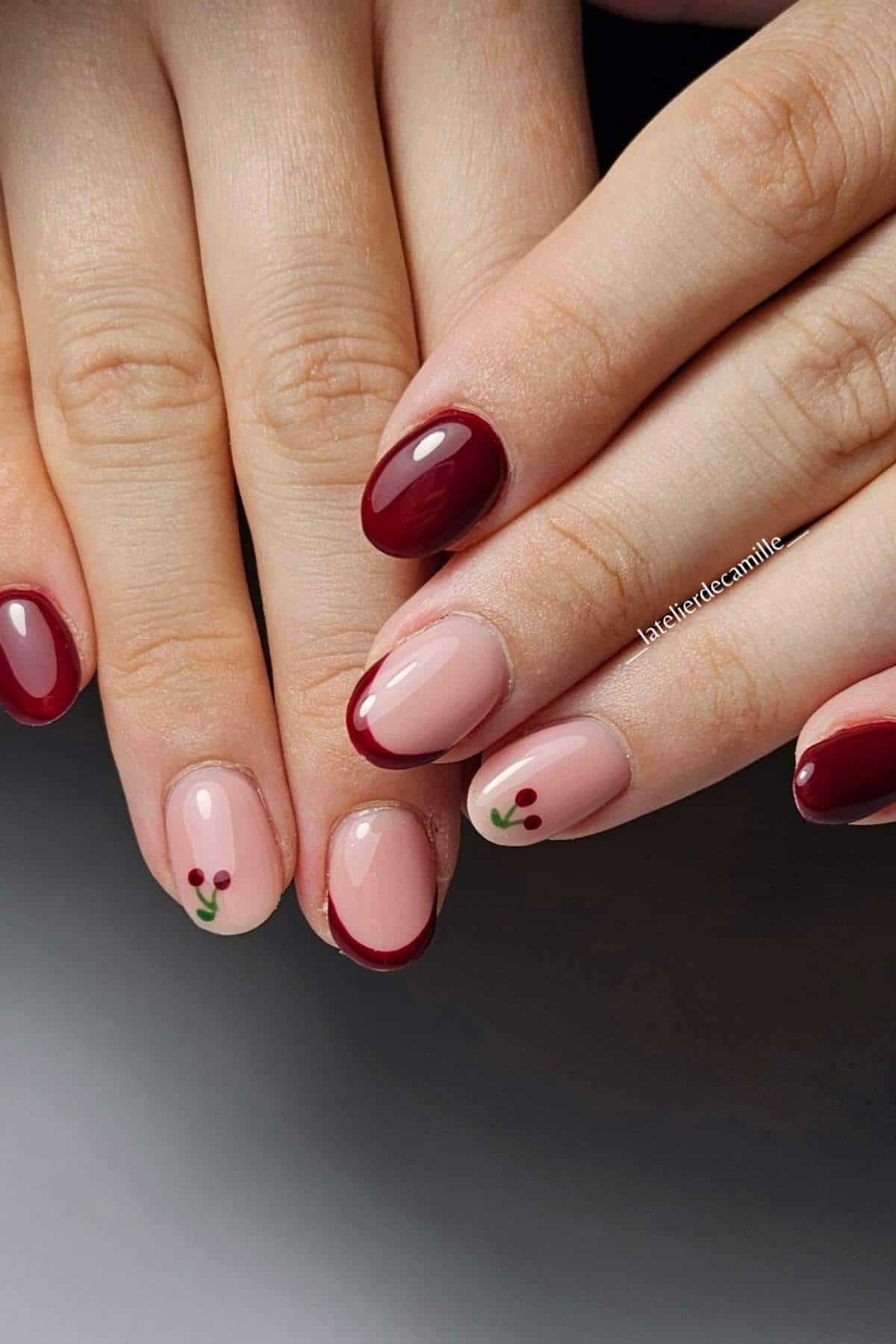 Hands with manicured nails featuring deep red polish, French tips, and small cherry designs on a light beige background.