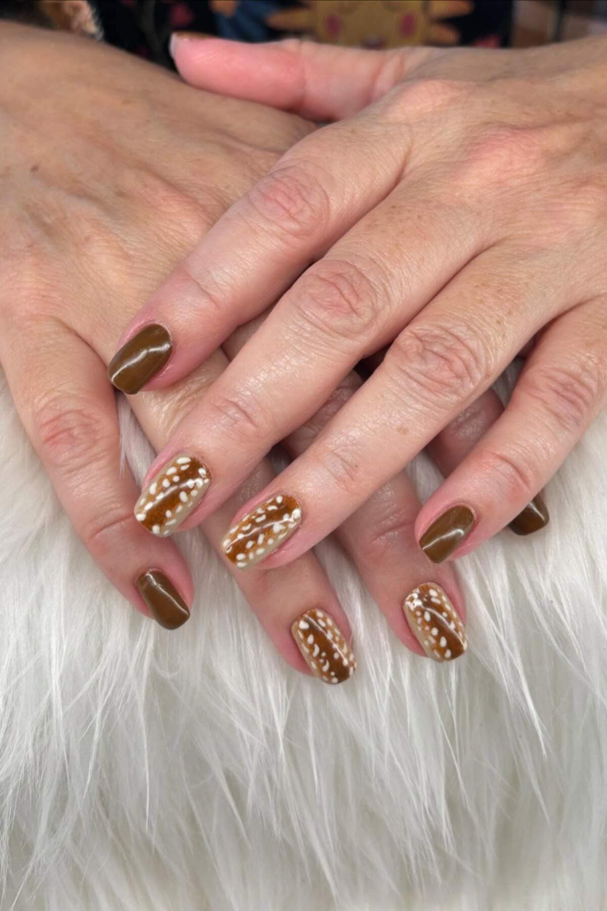 Hands with brown nail polish and white dotted designs on ring and middle fingernails, resting on a white furry surface.