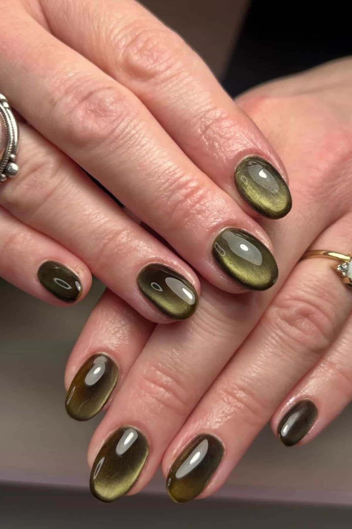 Close-up of hands with oval-shaped nails painted in a glossy olive-green cat-eye design, showing a gradient effect and reflective surface. Fingers are adorned with silver and gold rings.