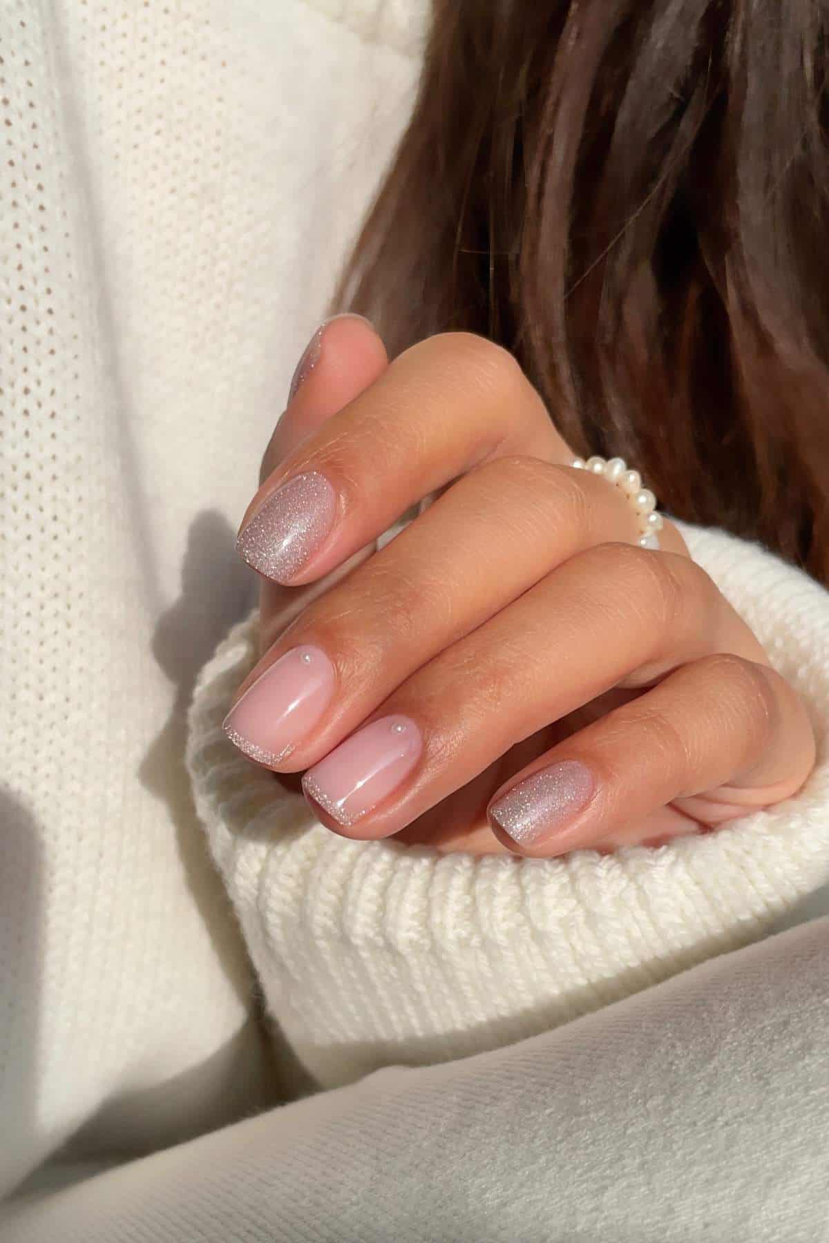 A hand with short, neatly manicured nails featuring a natural pink base and subtle silver glitter tips, resting on a white knitted sleeve.