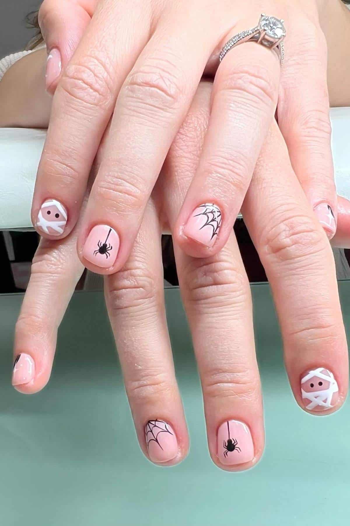 Hands with pale pink Halloween-themed nail art featuring ghosts, spiders, spider webs, and mummy designs. Person wears a diamond ring on the ring finger.