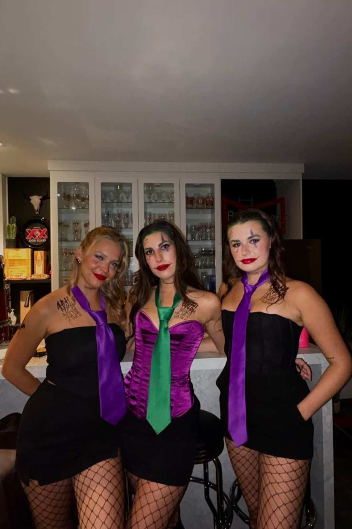 Three women pose indoors wearing black dresses, fishnet stockings, colorful ties, and Joker-inspired makeup, standing in front of a bar cabinet.