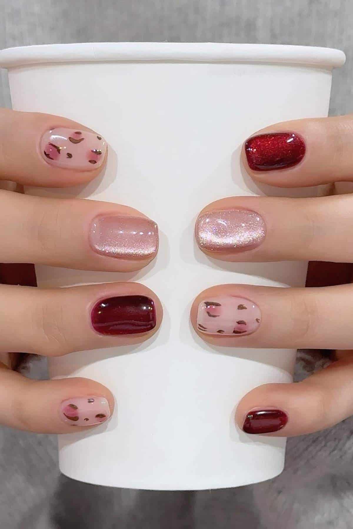 Hands holding a white paper cup, nails painted with alternating burgundy, pink glitter, and nude polish with small burgundy spots.
