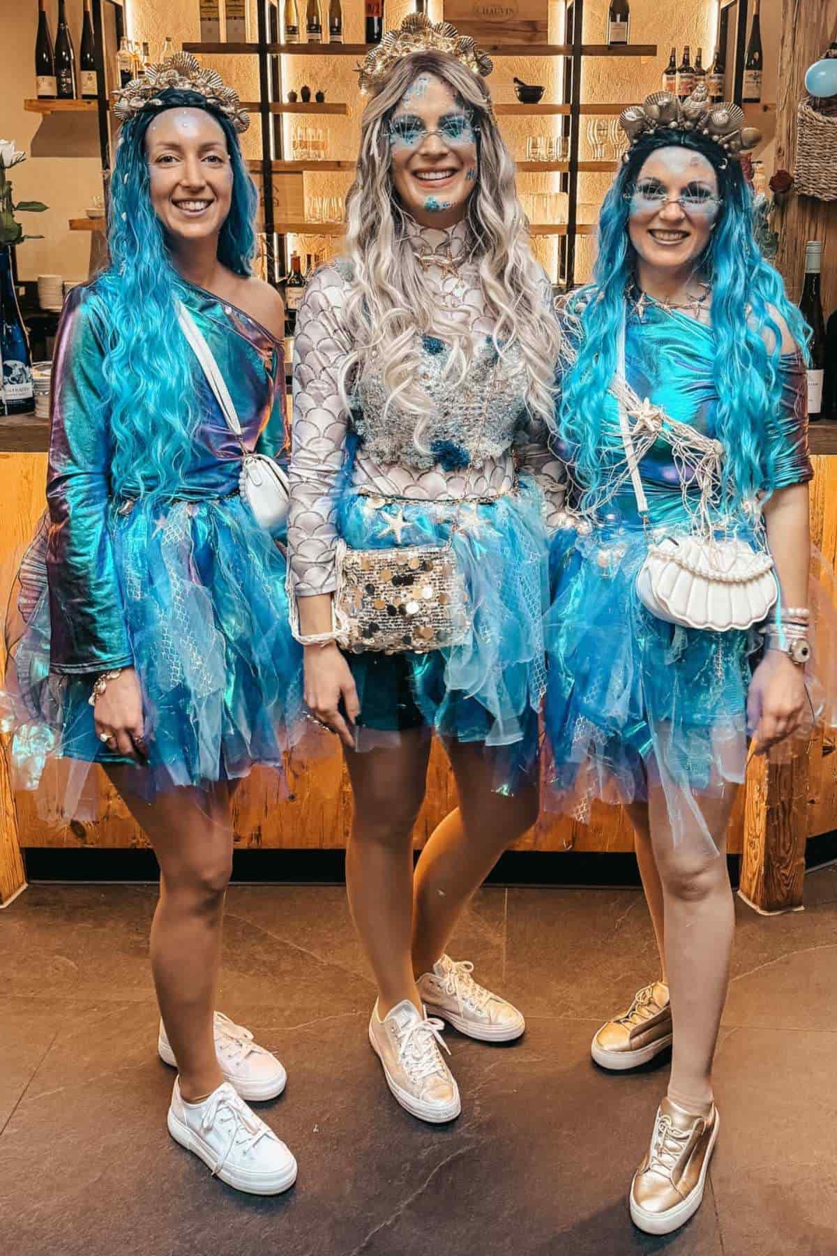 Three people stand indoors wearing matching blue and silver mermaid costumes, with blue wigs, sparkly makeup, and sneakers, posing and smiling for the camera.