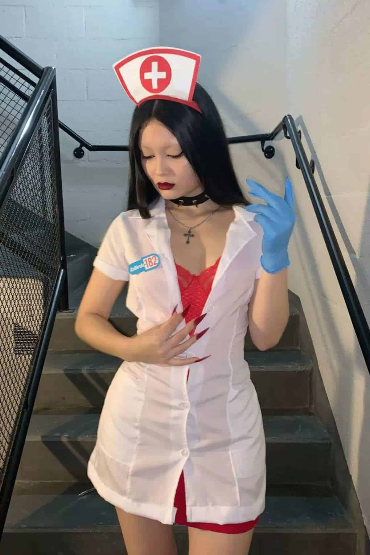 Person dressed in a nurse costume with a white dress, red lace top, nurse hat, black choker, and blue glove, standing on stairs indoors.